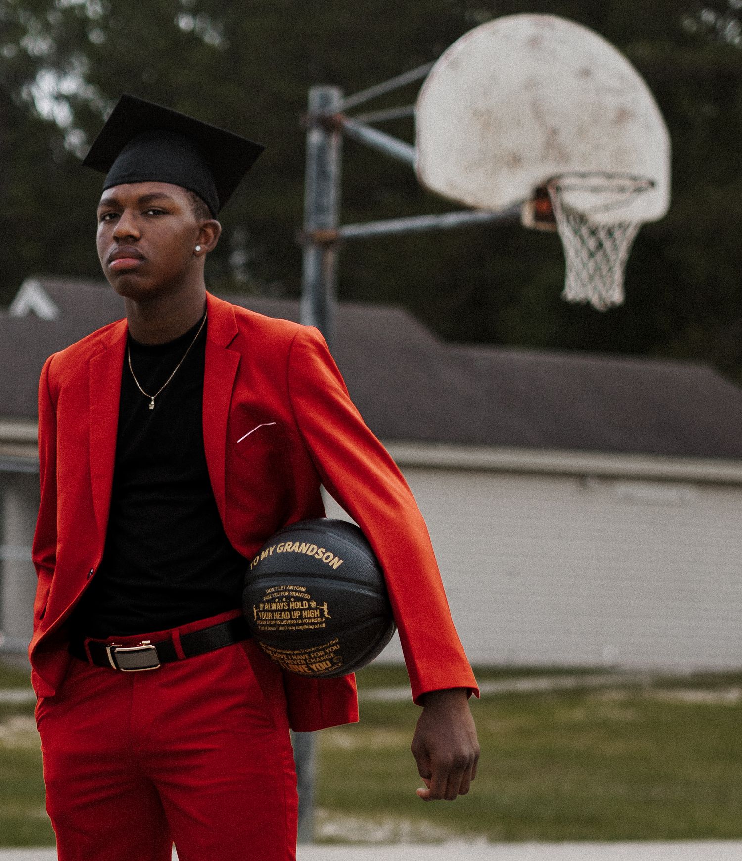Graduate in bright red suit jacket poses with basketball near outdoor court at dusk.