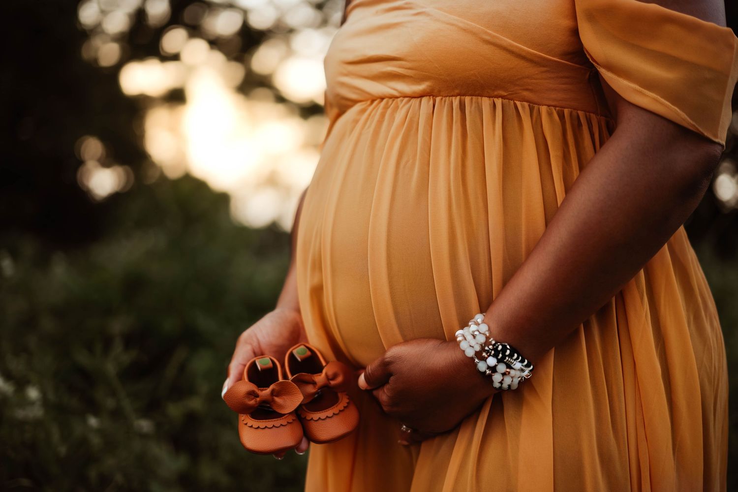 Expectant parent holding tiny orange baby shoes against sunset backdrop.