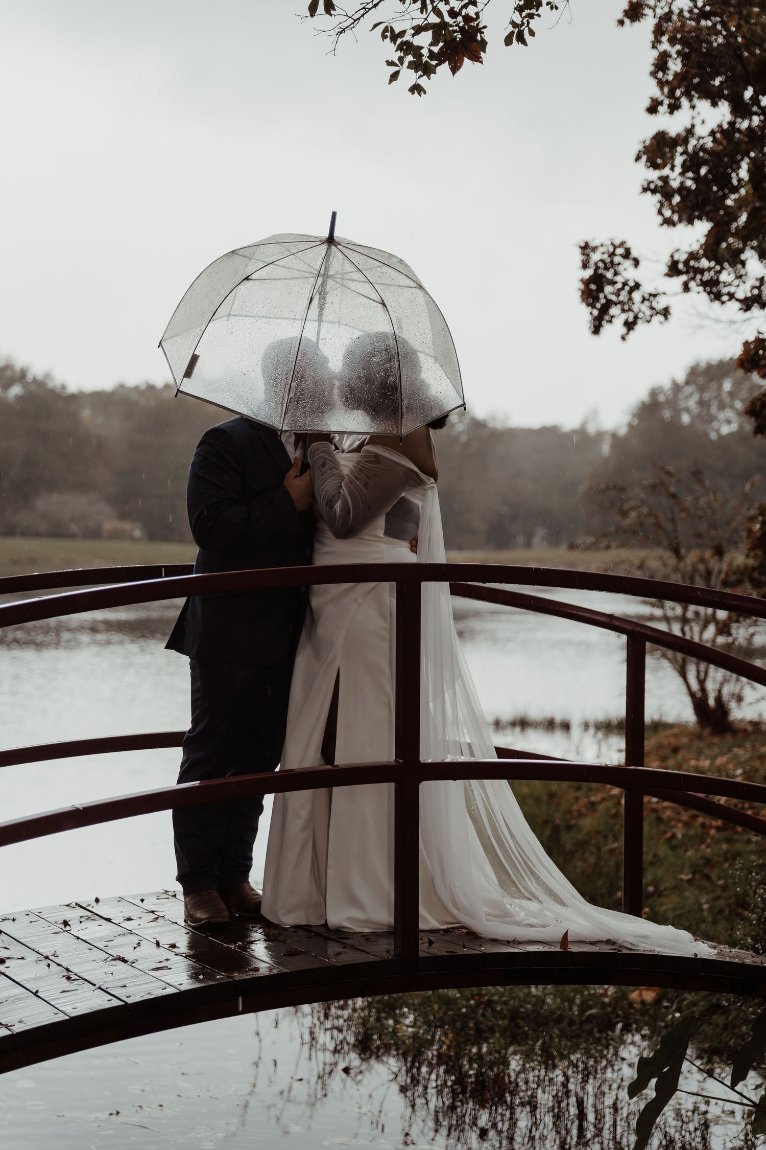 A romantic rainy day wedding photo on a bridge with a clear umbrella.