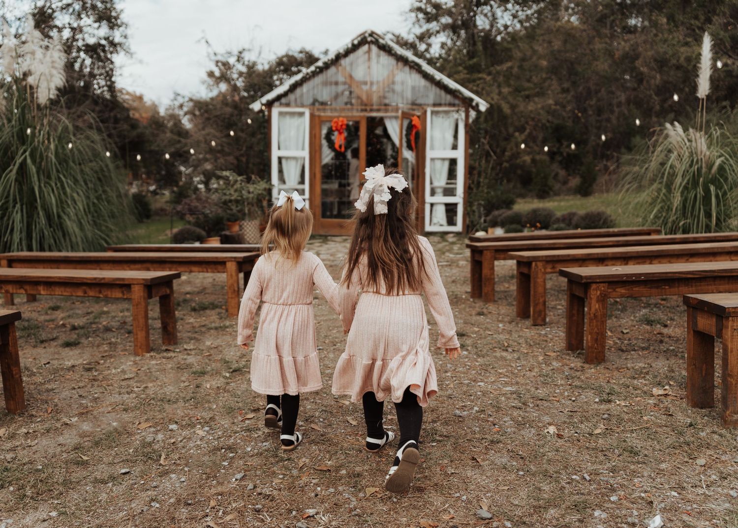 Two children in pink dresses walk toward rustic shed with wooden benches.