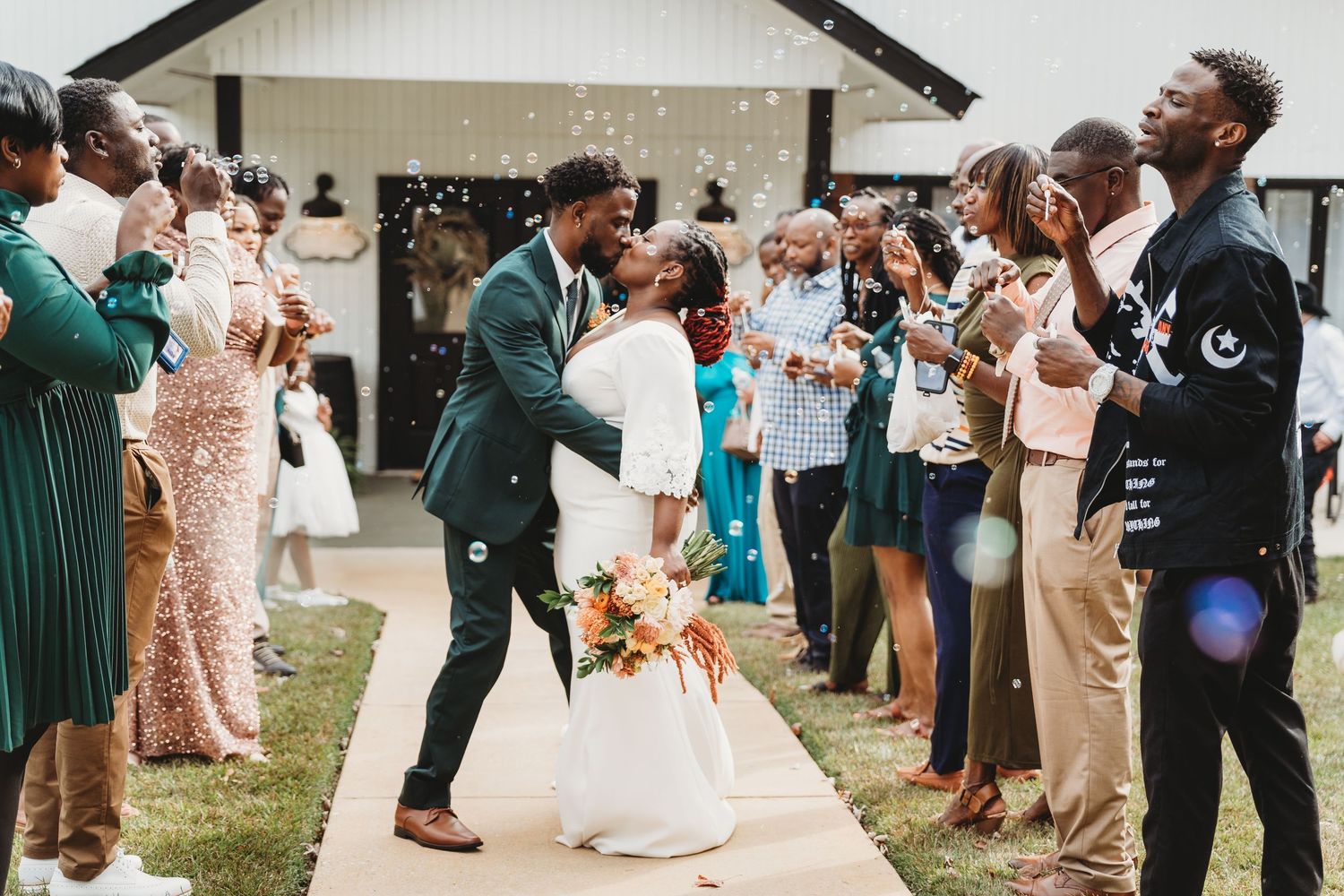 Wedding guests form aisle for newlyweds during outdoor celebration near white building.
