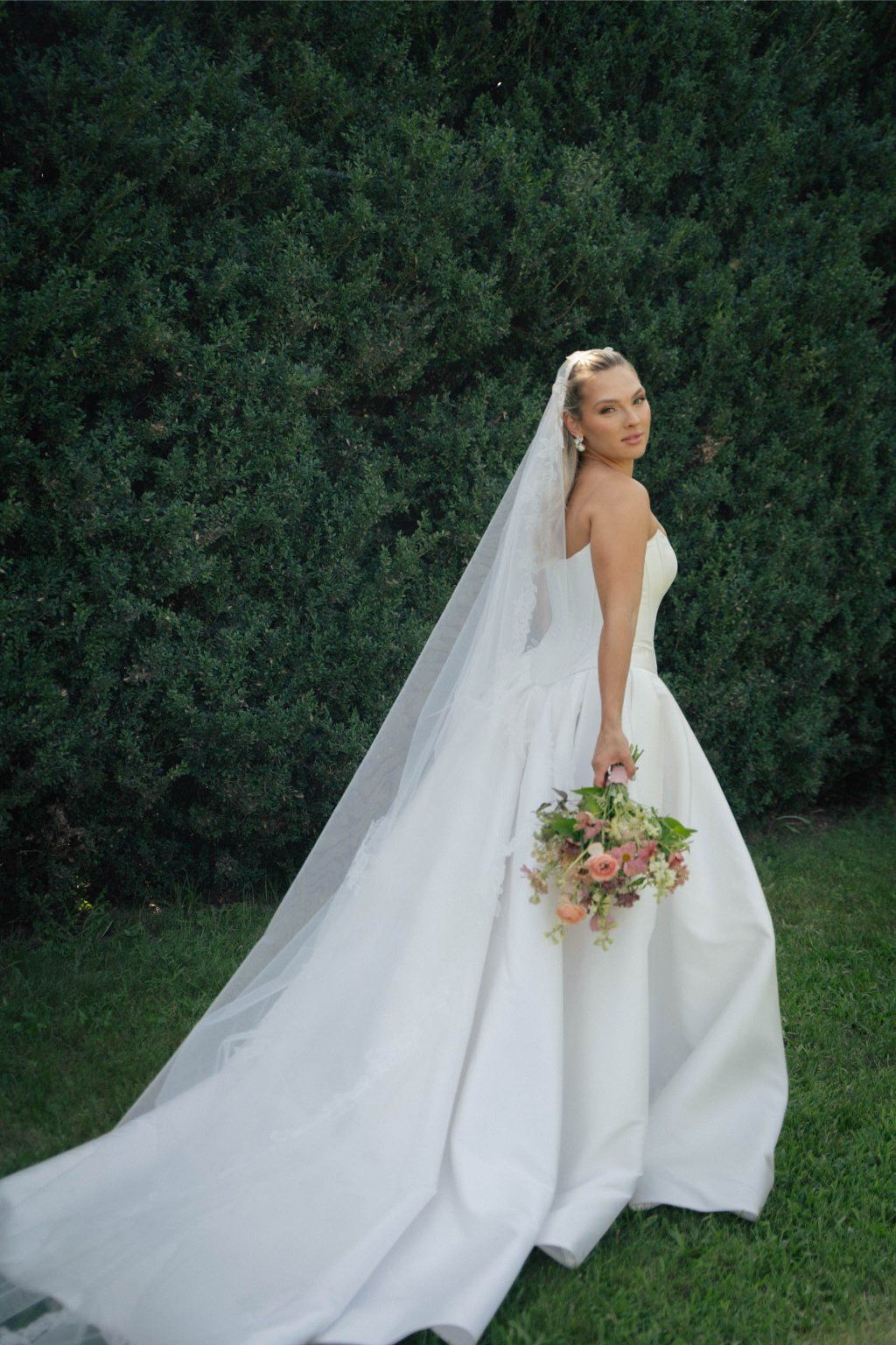 A bride in a flowing white wedding dress with a long train poses elegantly against a lush green garden backdrop.
