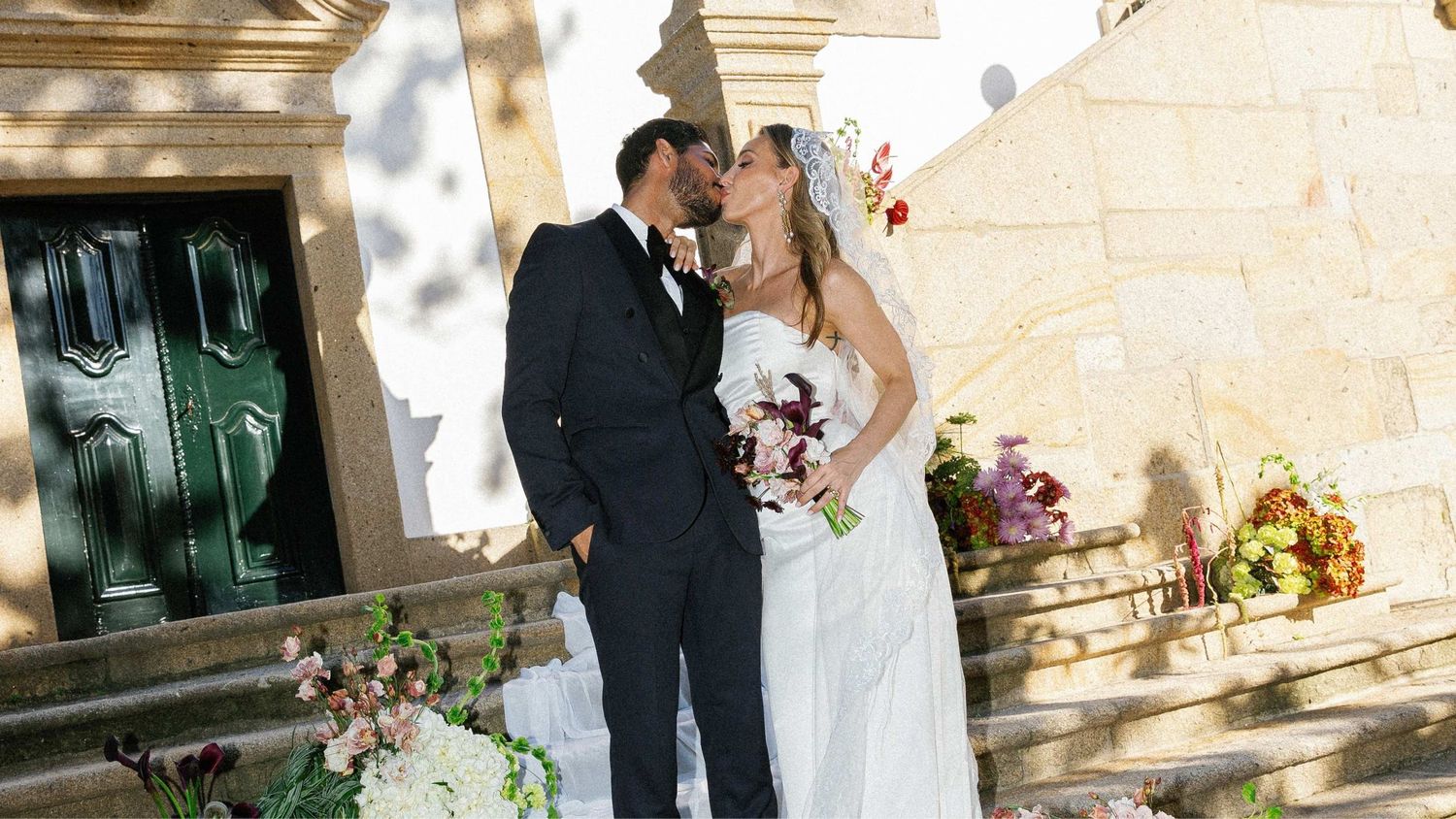 A couple in wedding attire share an intimate moment on a sun-dappled stone patio with floral decorations.
