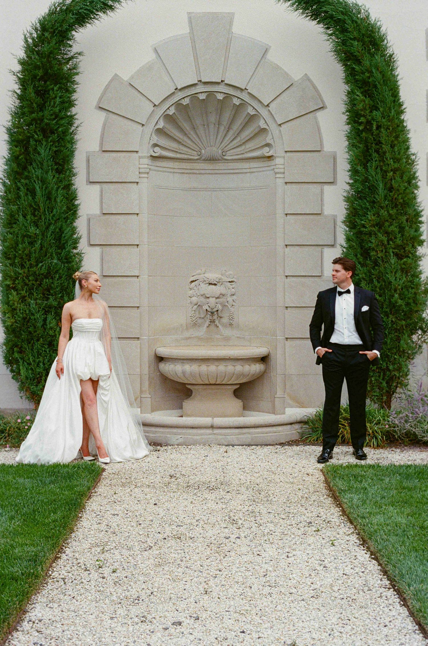 A bride and groom pose for creative wedding photos against a stone wall with fountain and cypress trees.