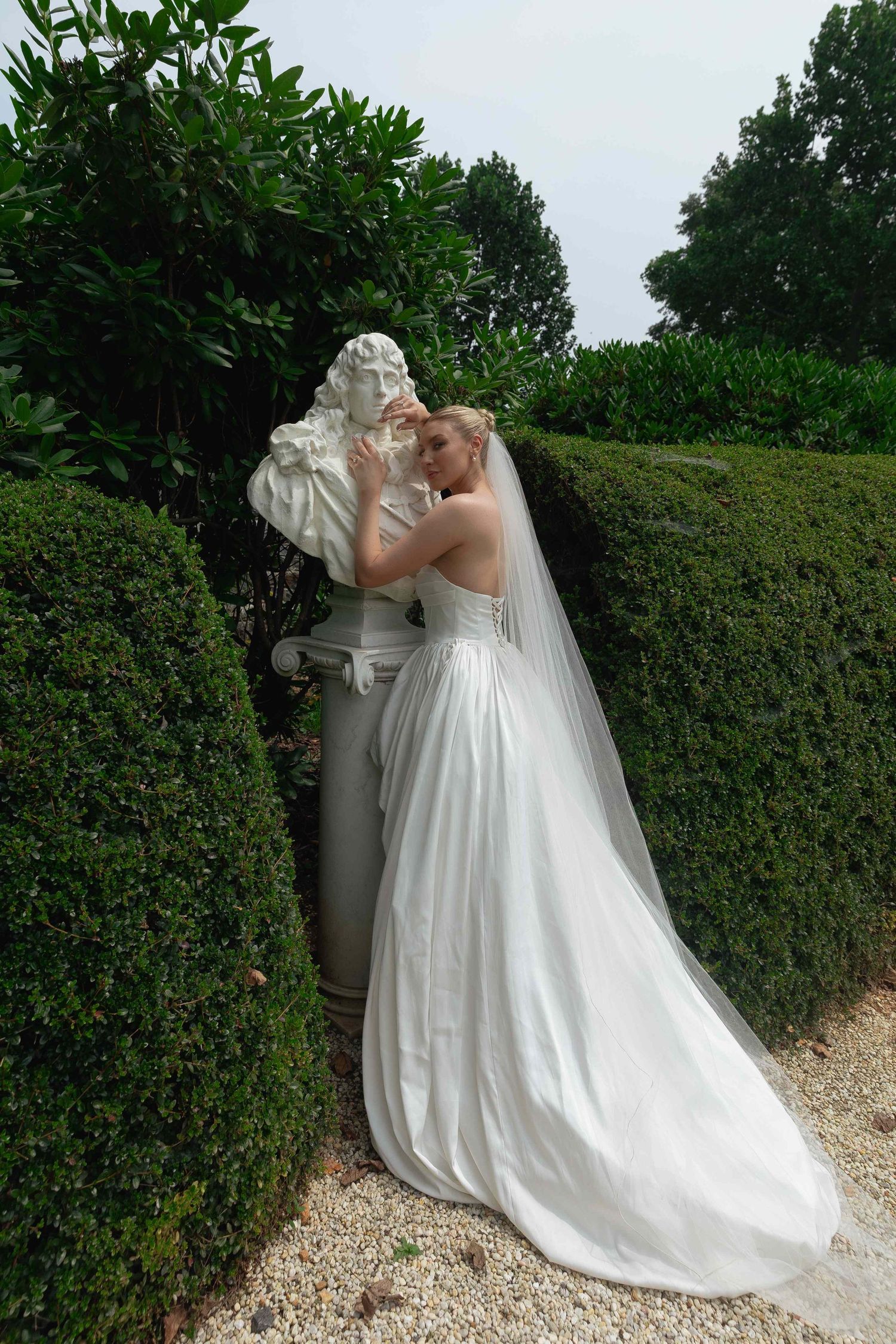 A couple in wedding attire share a romantic moment along a hedge-lined garden path.