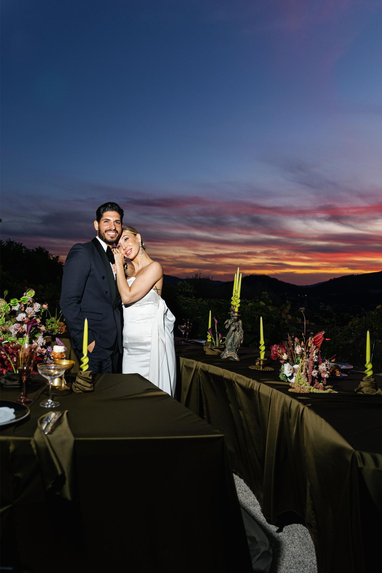 A couple embraces on an outdoor terrace during a stunning sunset with vibrant pink and orange skies.