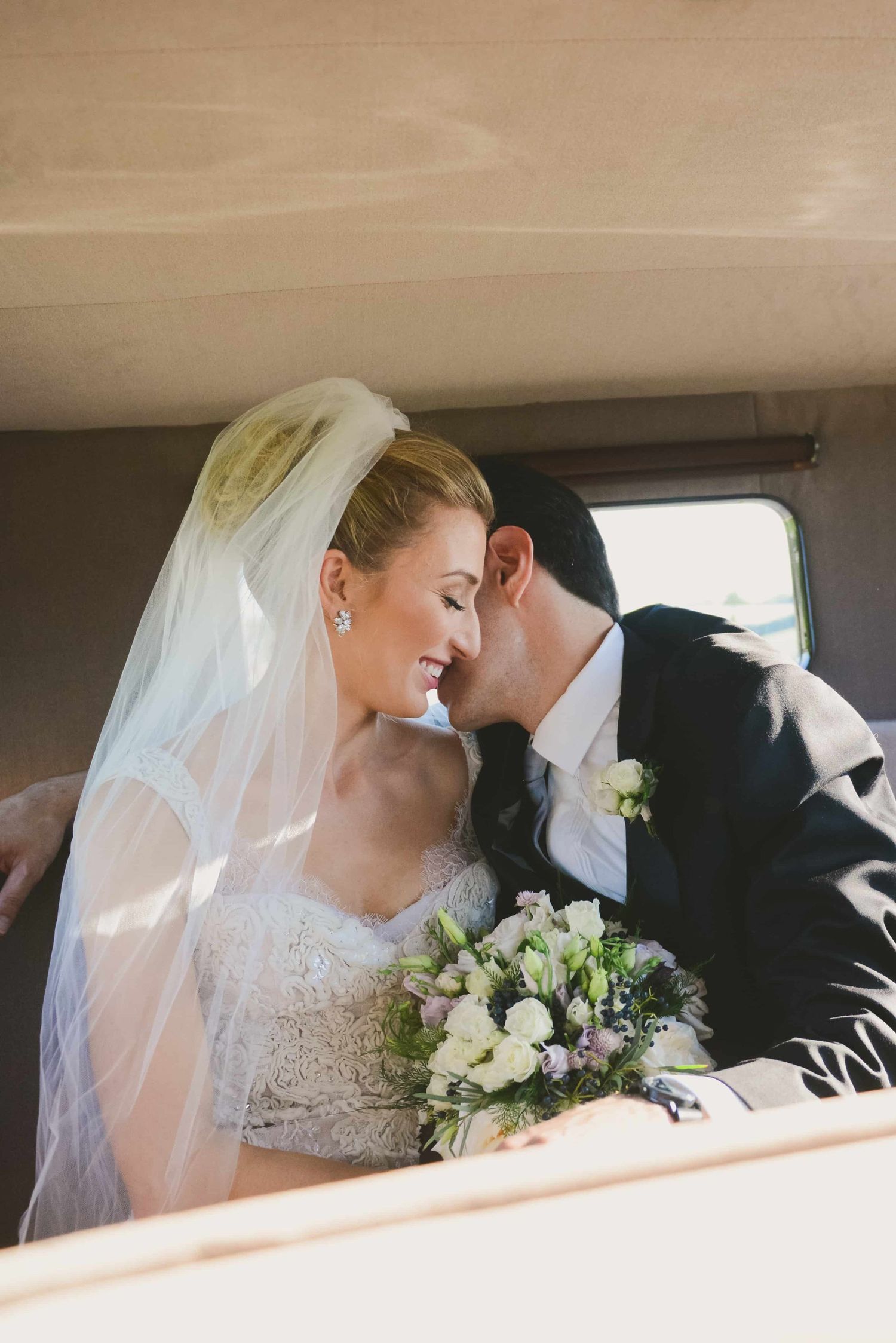 A romantic wedding couple shares an intimate moment in the backseat of a vintage car while holding a white floral bouquet.