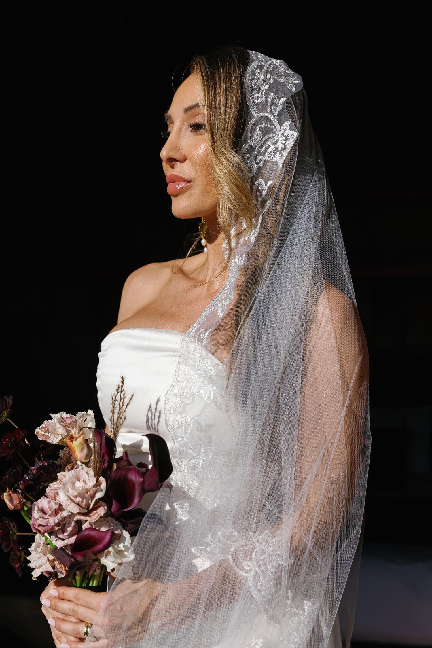 A bride in a white wedding dress and delicate lace veil poses with a pink and burgundy floral bouquet against a dark background.