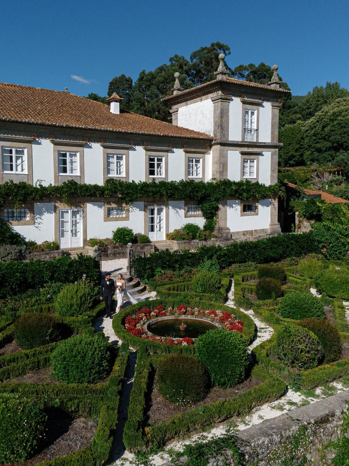 Traditional white Portuguese houses with blue trim nestled on a hillside garden with manicured hedges and red flowers.