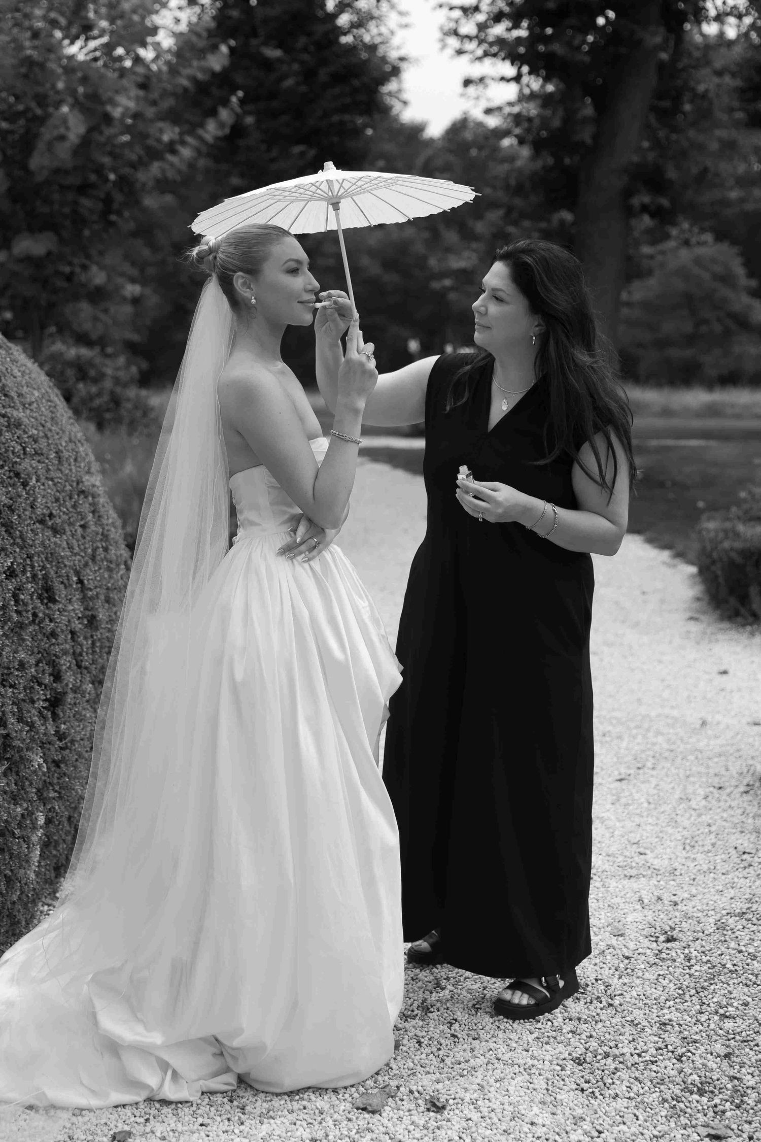 Two figures sit on a gravel path sharing a parasol in black and white photography.