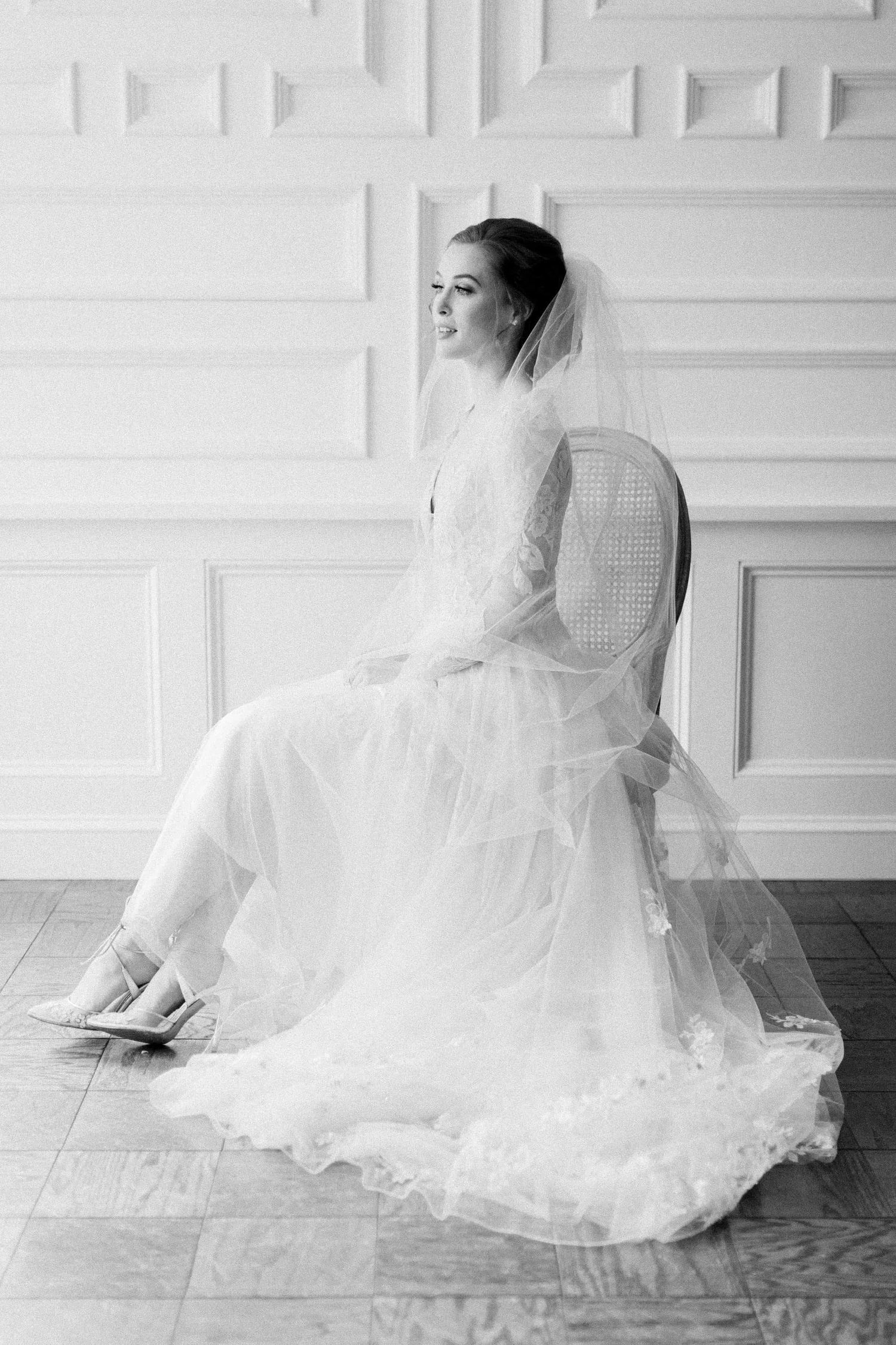 A bridal gown with delicate lace details and flowing tulle skirt creates an ethereal silhouette against a white textured wall.