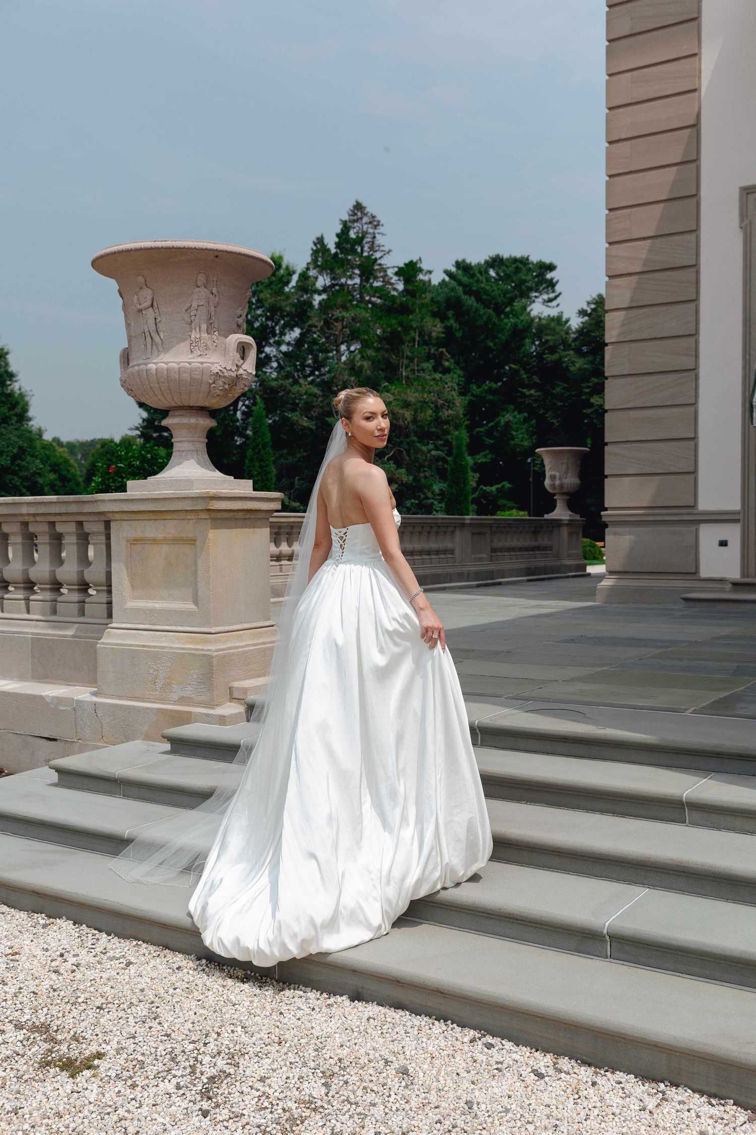 A bride in a flowing white ballgown walks across an elegant stone terrace with decorative urns.