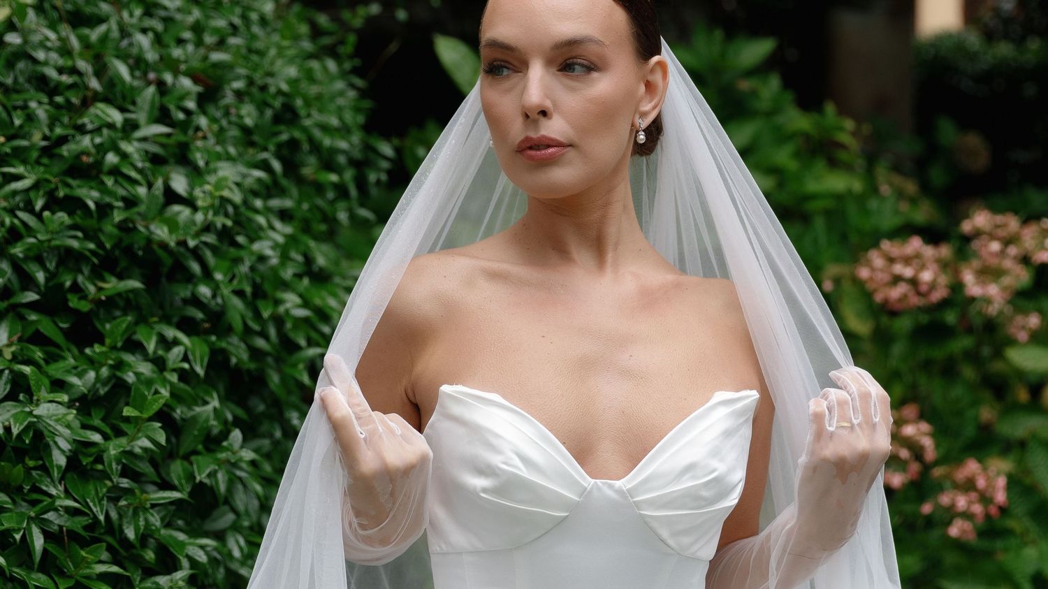 A bride in a strapless white wedding gown stands elegantly against greenery in an outdoor setting.