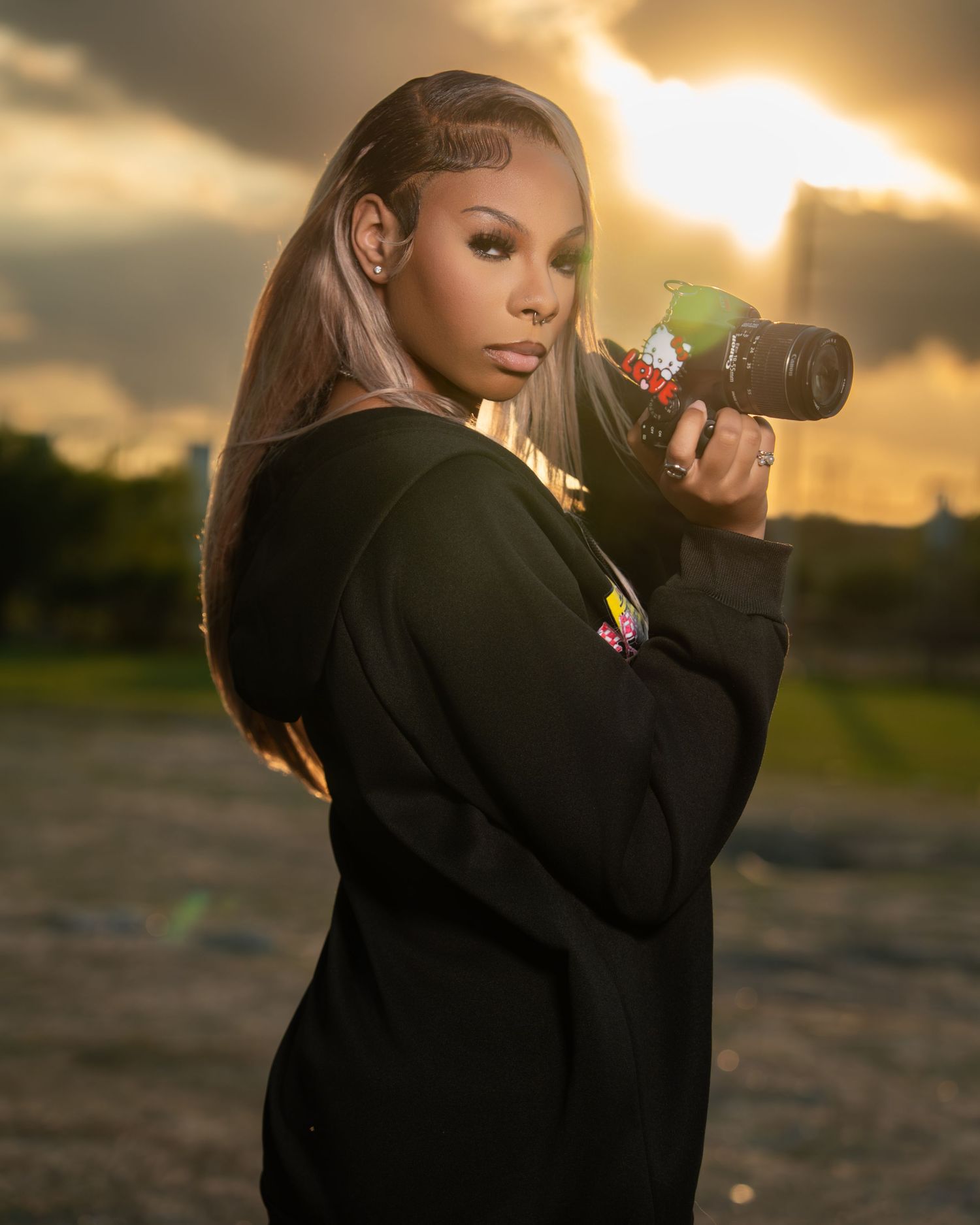 Woman photographer poses with camera at golden hour sunset, wearing black hoodie with long straight hair.
