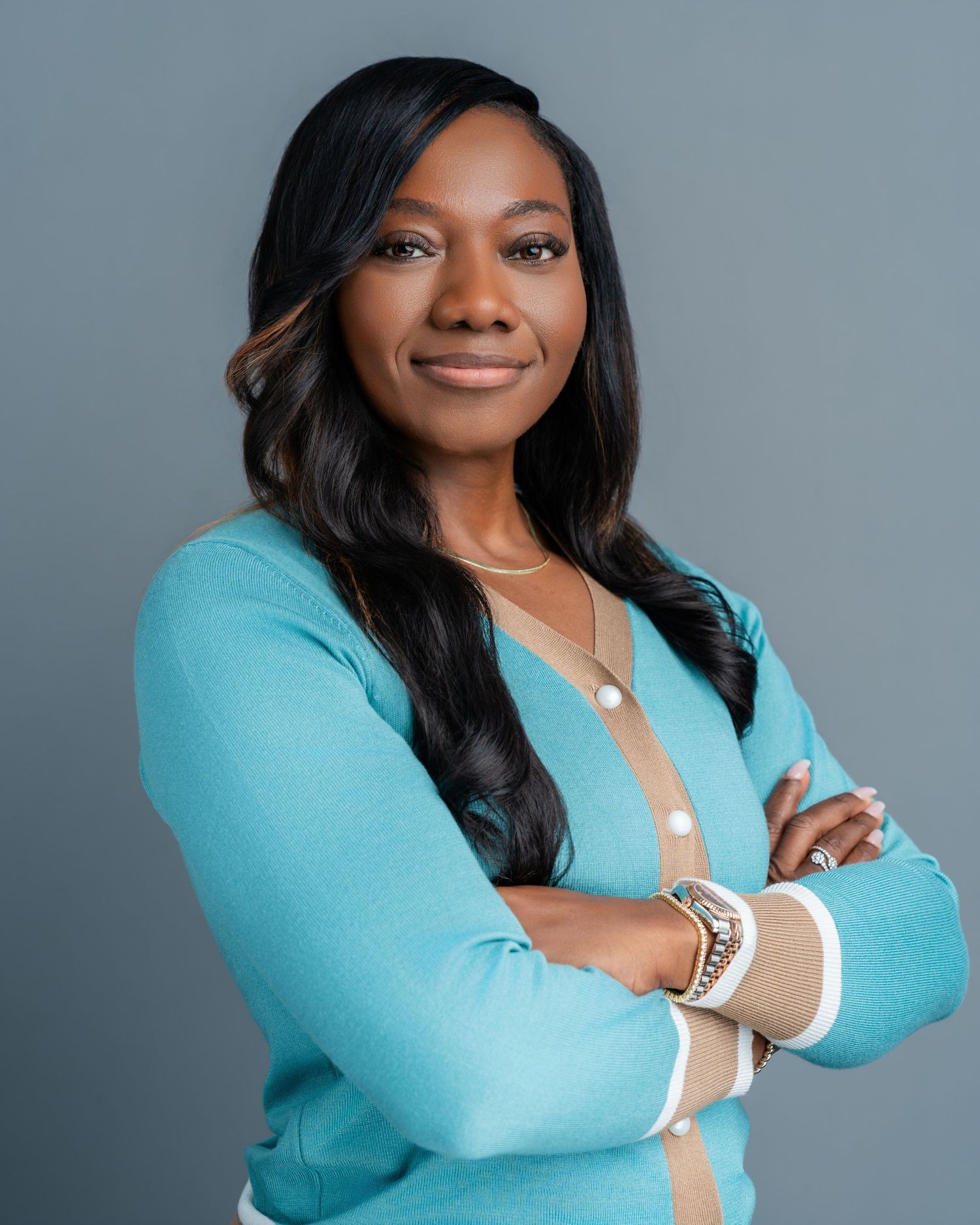 Professional headshot of a confident woman in a teal cardigan with arms crossed, smiling against a gray background.