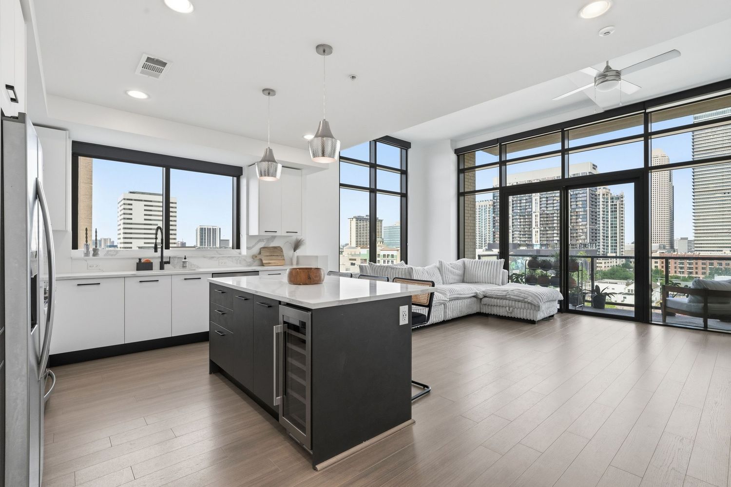 Modern open-concept kitchen with island, wine fridge, white cabinets, and floor-to-ceiling windows with city views.