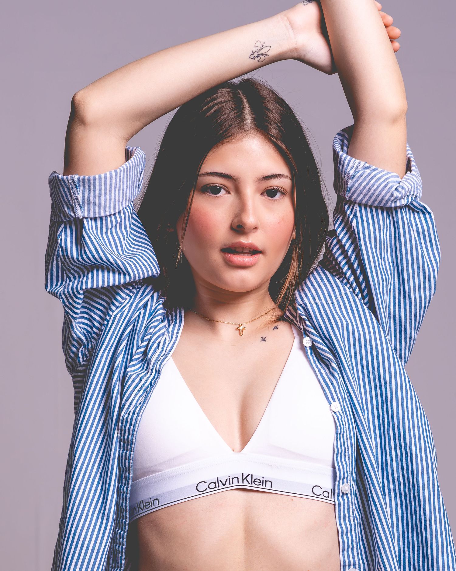 Young woman in Calvin Klein bralette and blue striped shirt posing with arms raised against gray background.