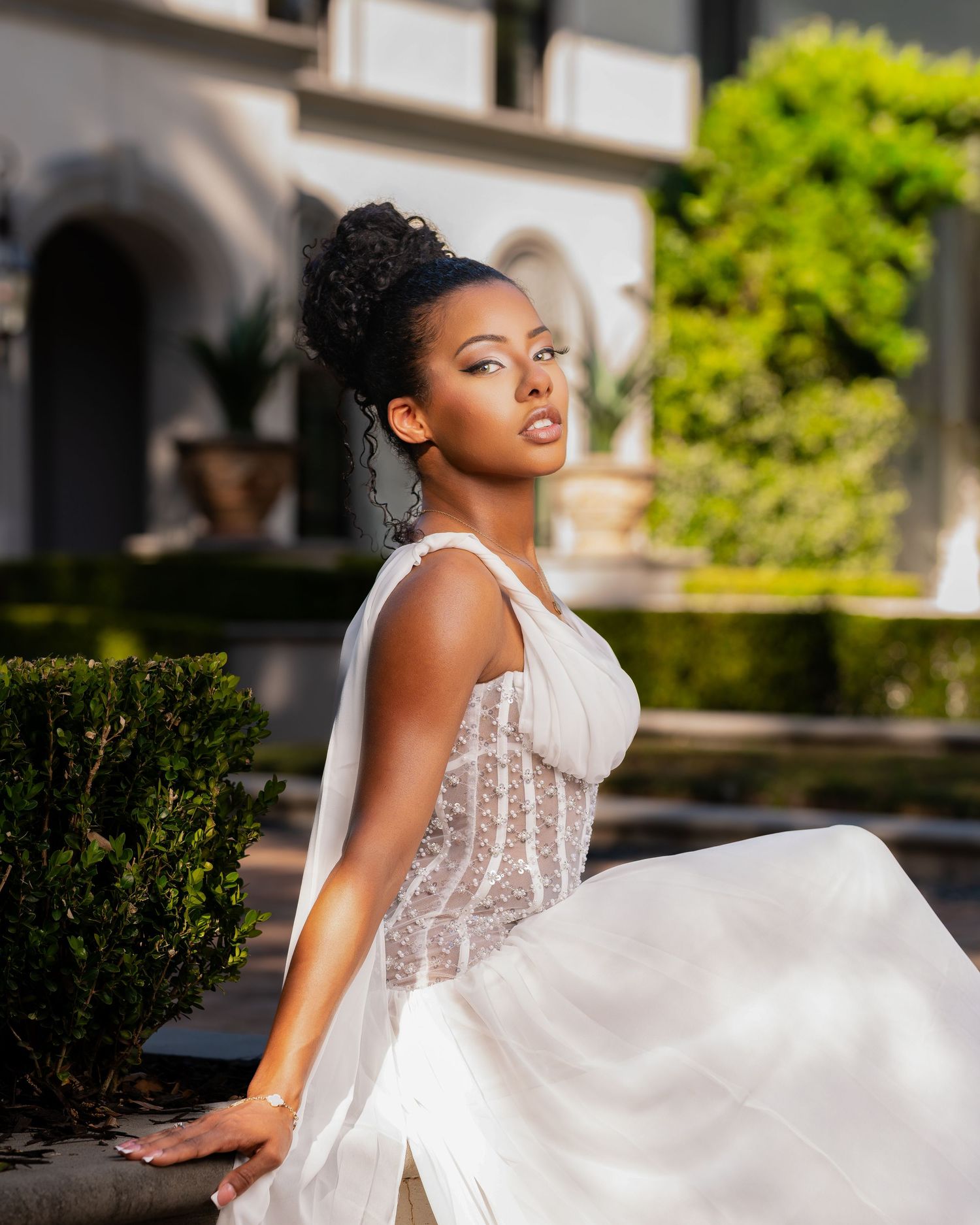 Elegant woman in white lace gown poses gracefully in a lush garden setting with classical architecture.