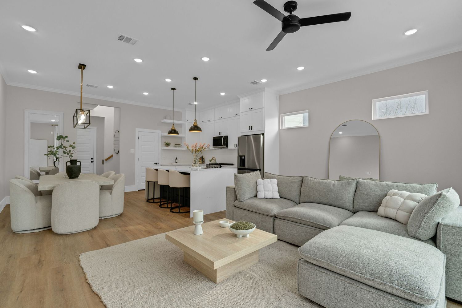 Modern open-concept living room with gray sectional sofa, light wood floors, and white kitchen with pendant lights.