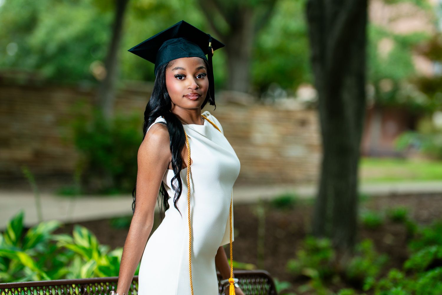 Graduate in white dress poses outdoors with graduation cap in garden setting. Powerrpackmedia Houston Texas