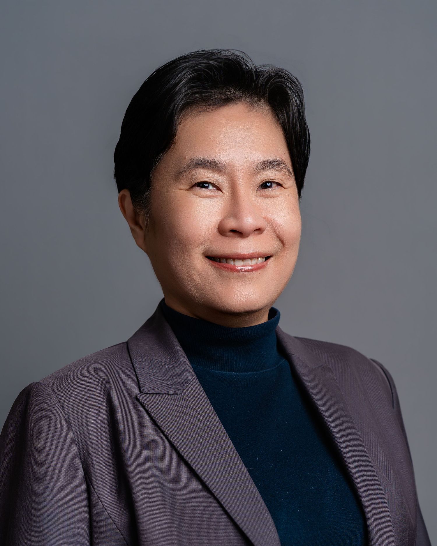 Professional headshot of an East Asian person in a gray blazer and teal turtleneck, smiling confidently against a gray background.