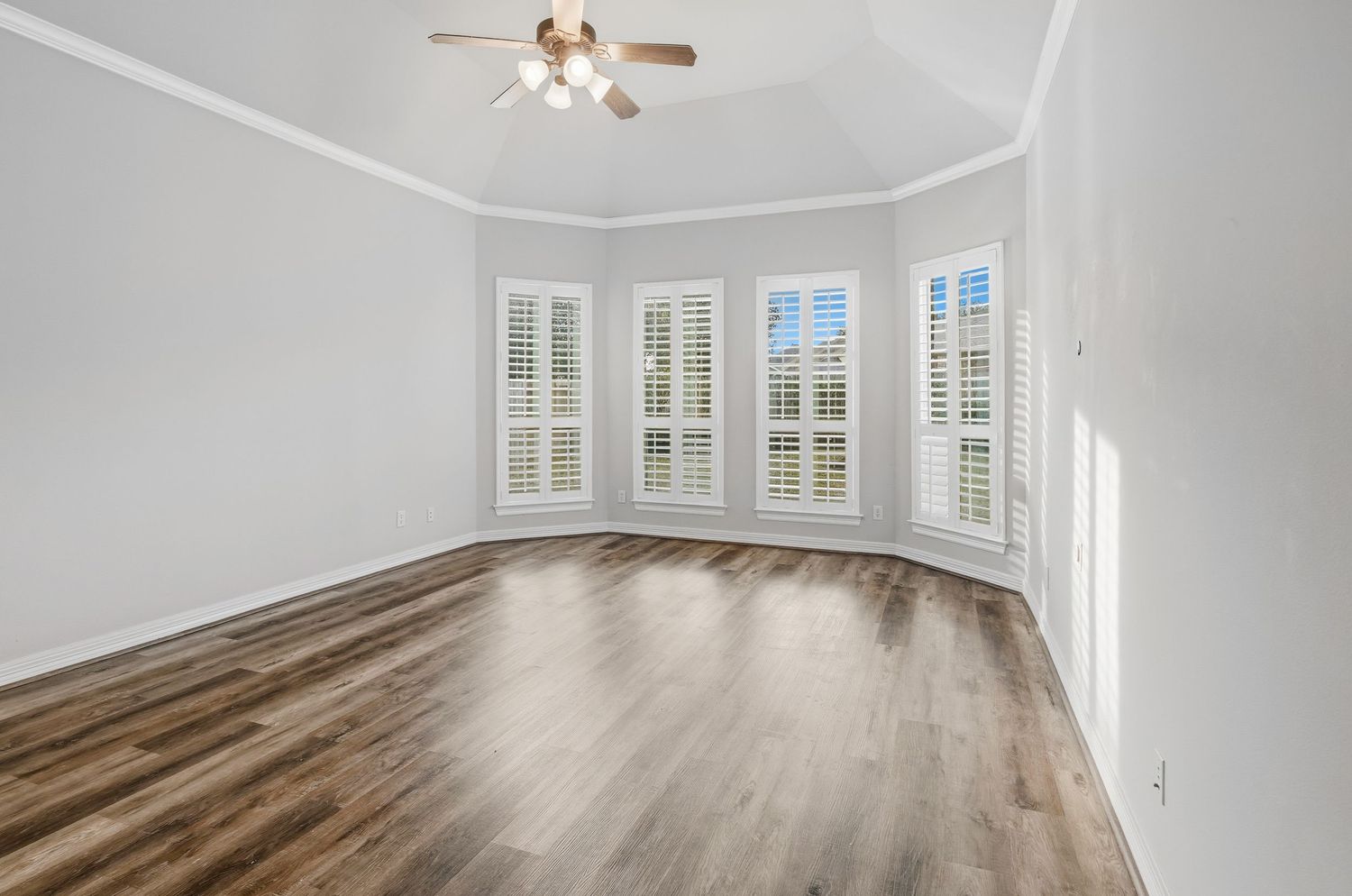 Empty room with bay windows, white plantation shutters, hardwood flooring, and a ceiling fan in natural lighting.