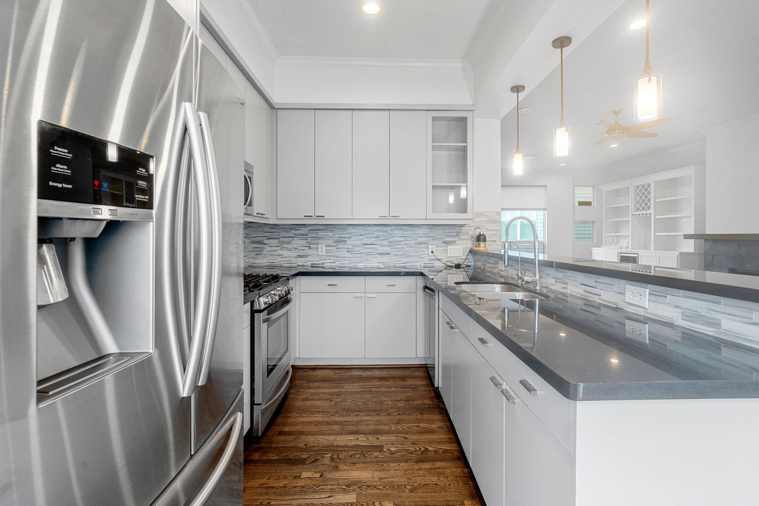 Modern white kitchen with stainless steel appliances, gray quartz countertops, pendant lights and hardwood flooring.