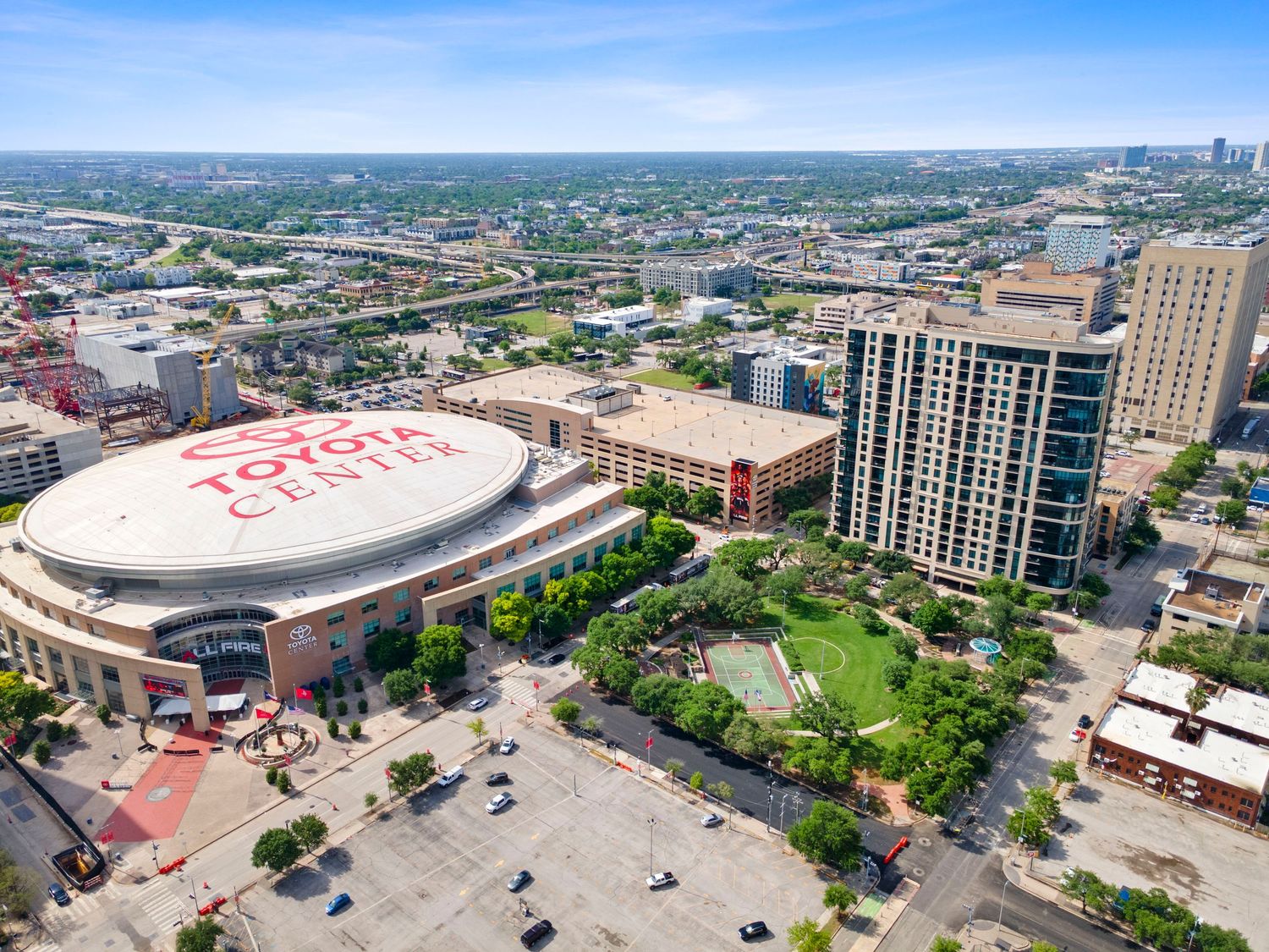 Aerial view of Toyota Center arena in downtown Houston, TX, surrounded by urban buildings and parking lots under a blue sky.