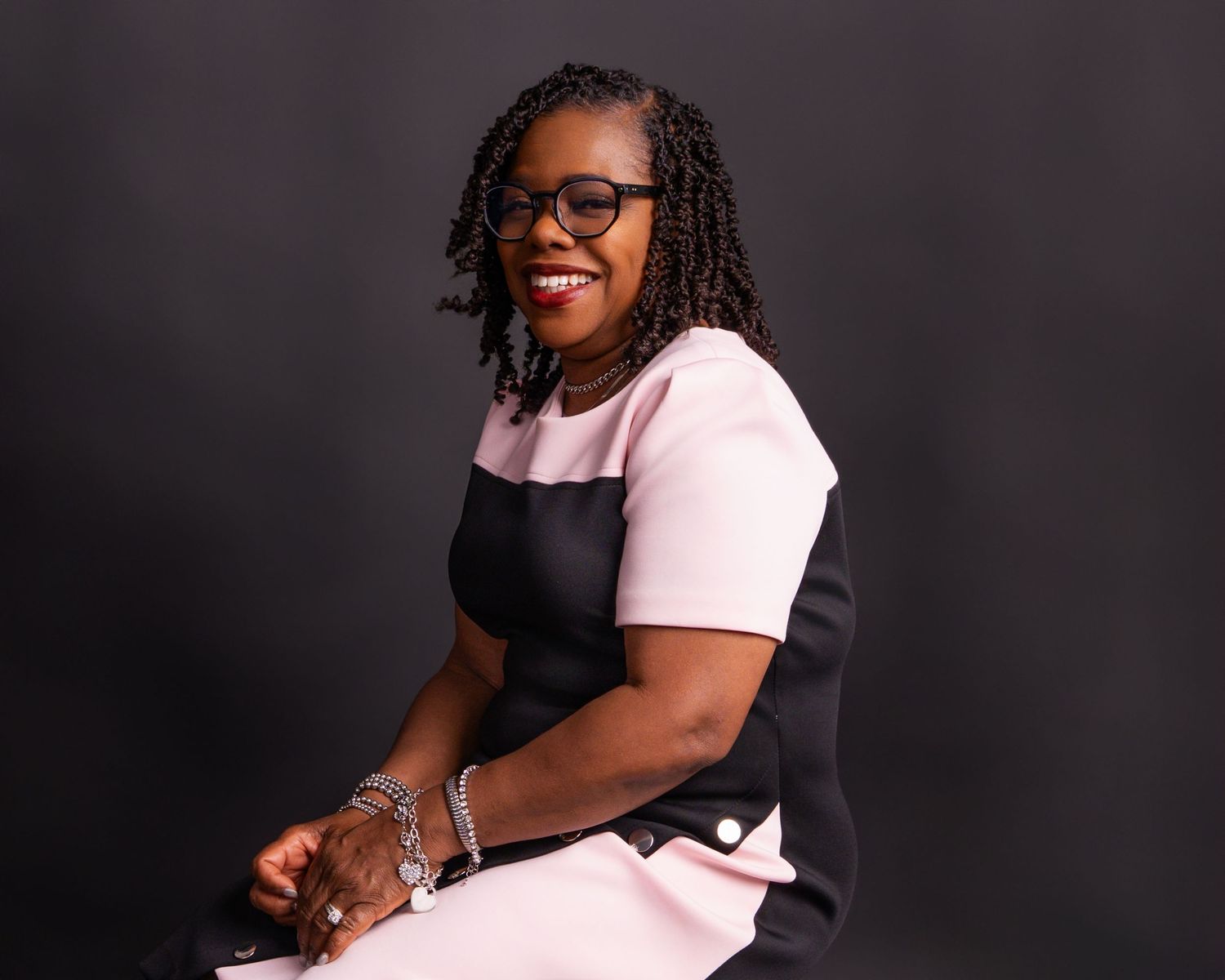 Professional woman smiling in pink and black dress, wearing glasses, seated against dark studio background.