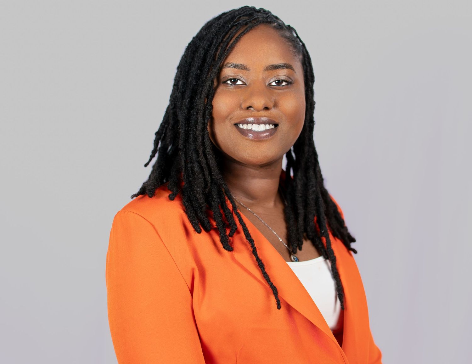 Professional headshot of someone wearing a bright orange blazer with a white top and dark hair against a gray background.