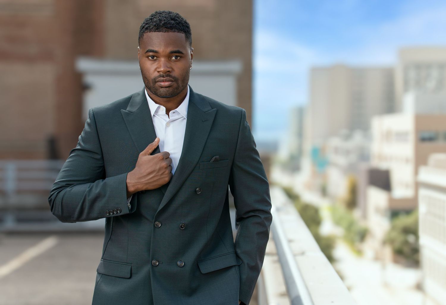 Professional in a dark green suit stands confidently on a rooftop terrace with city buildings in the background. Powerrpackmedia Houston Tex