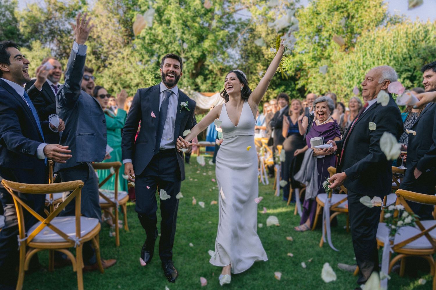 Wedding ceremony in progress on green lawn with wooden chairs and standing guests.