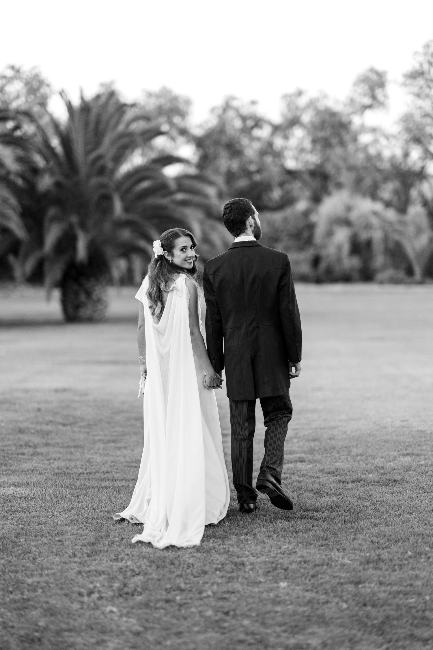 Black and white photo of newlyweds walking together across an expansive lawn.