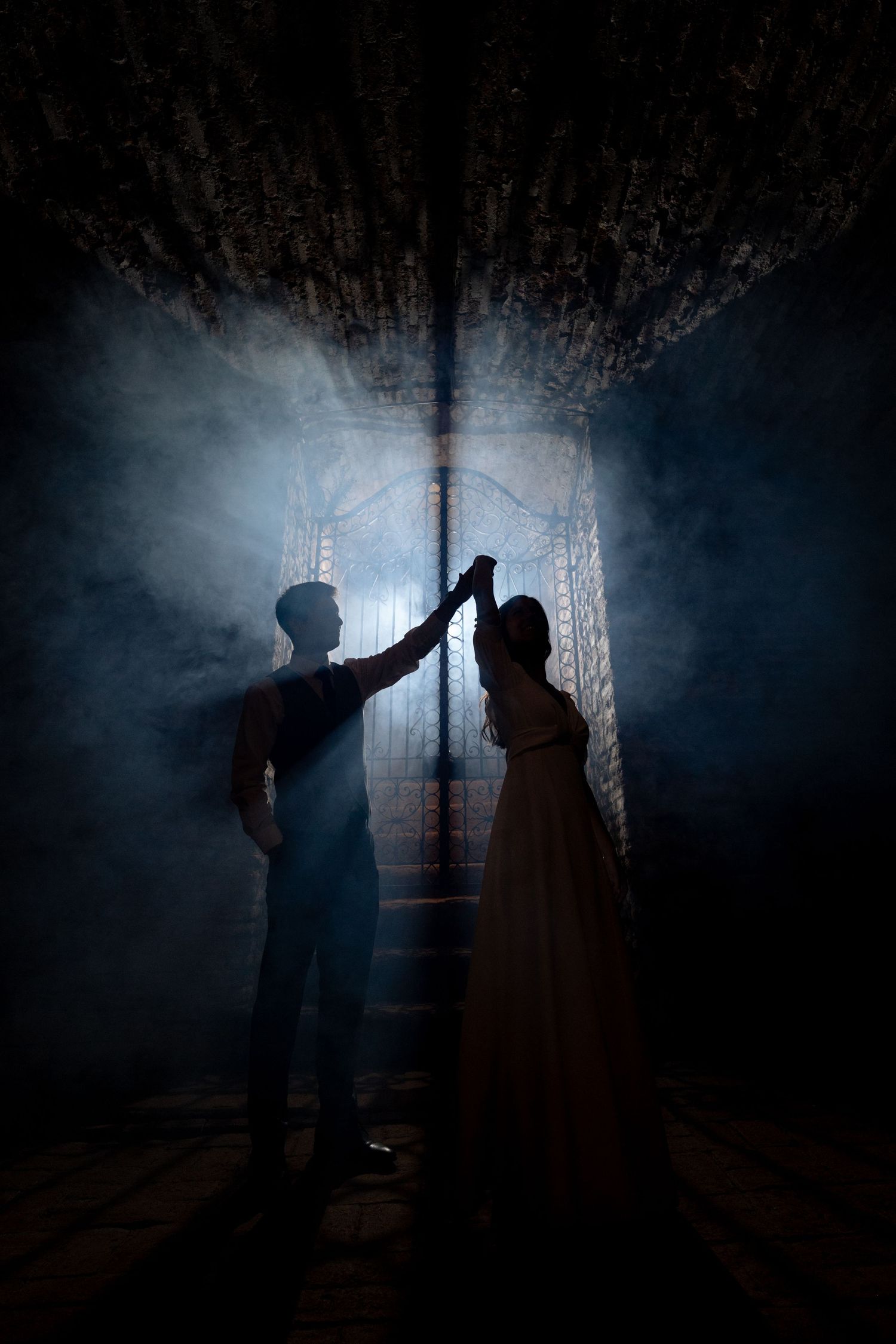 Silhouetted couple holding hands before ornate gate with dramatic blue smoky light in dark mysterious setting.