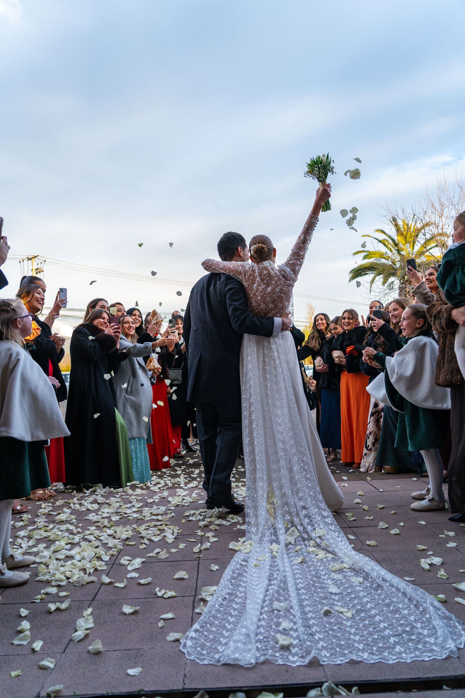 Bride in flowing lace train dress celebrates with guests throwing flower petals outdoors.