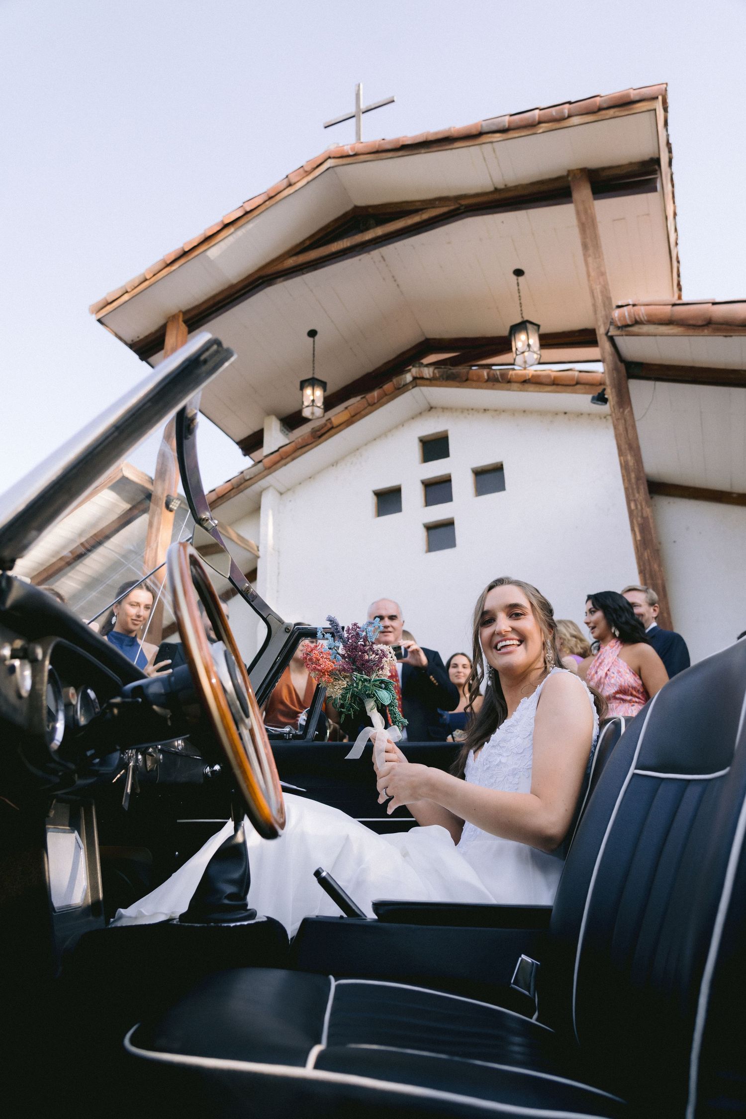 Wedding couple sitting in vintage convertible car outside white church building.