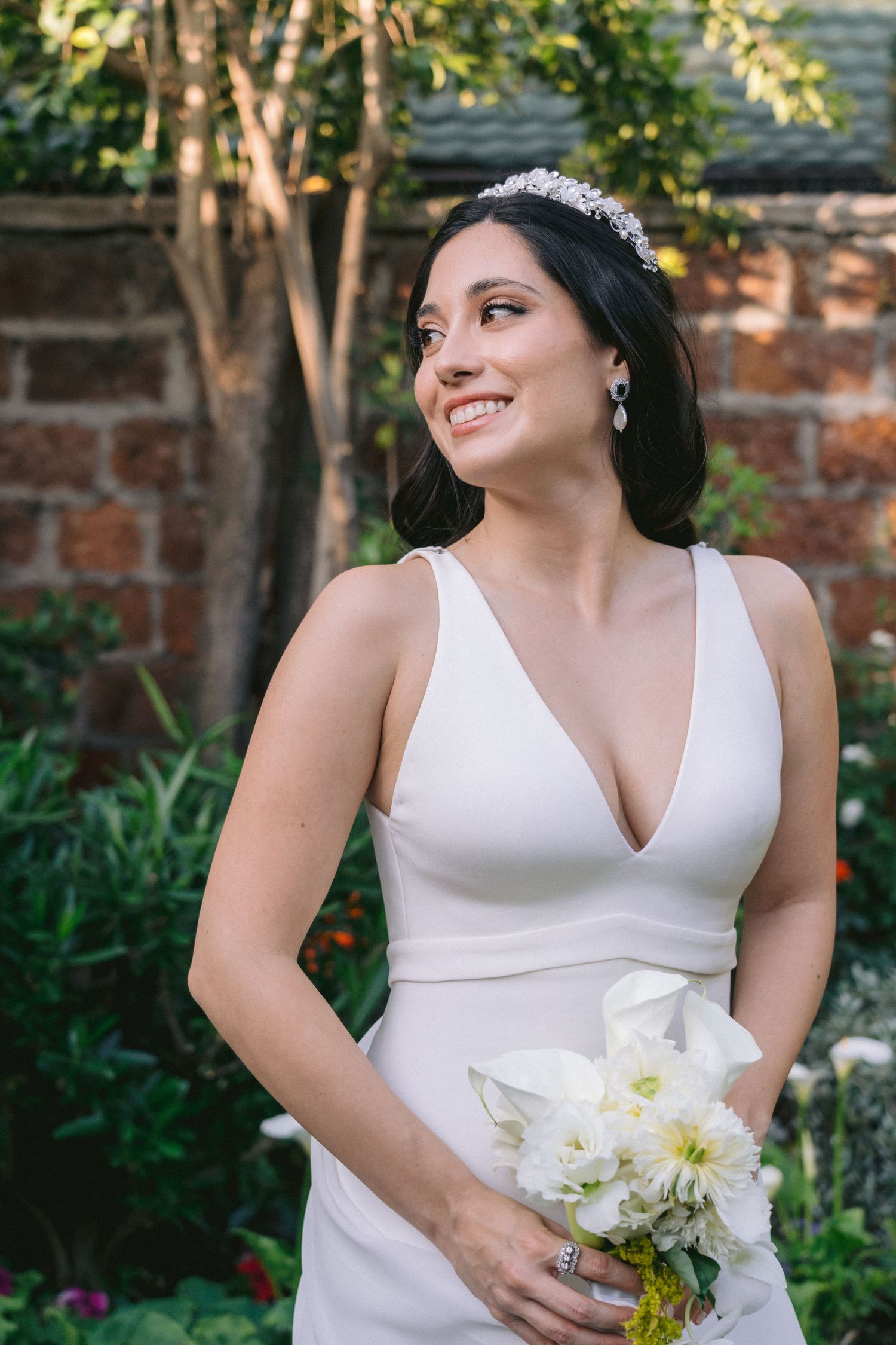 A bride in a classic white wedding dress poses in a garden setting holding a bouquet of white flowers.