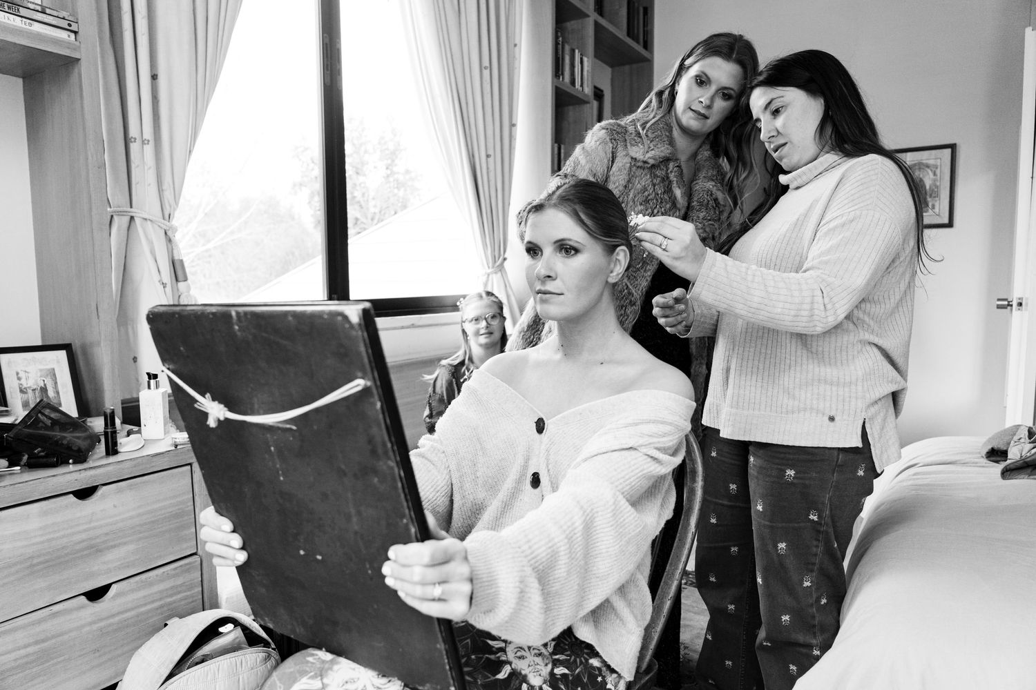 Wedding preparation scene showing stylists working on bridal hair and makeup in a bright room.