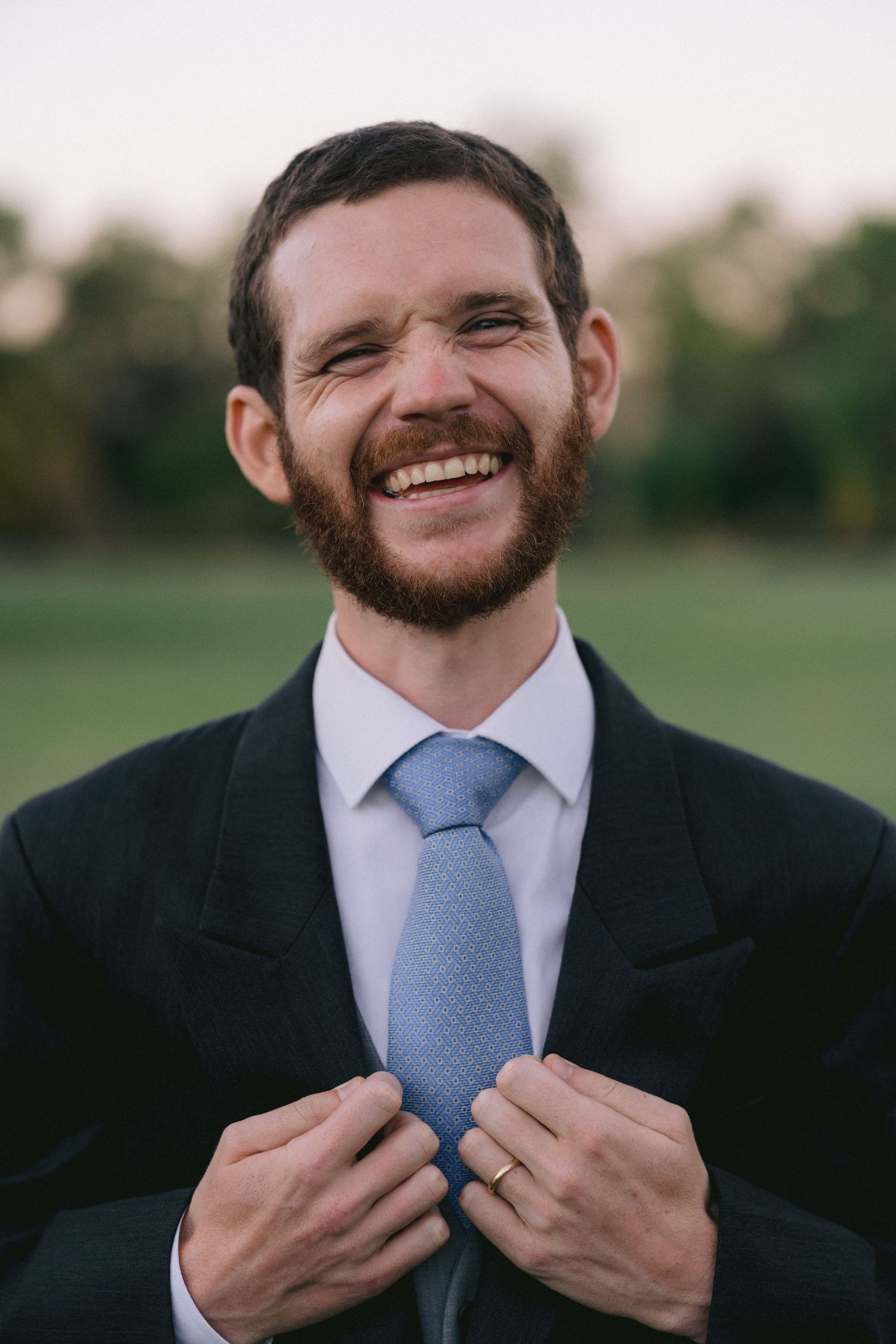 Person in formal suit with beard adjusting light blue tie while smiling.