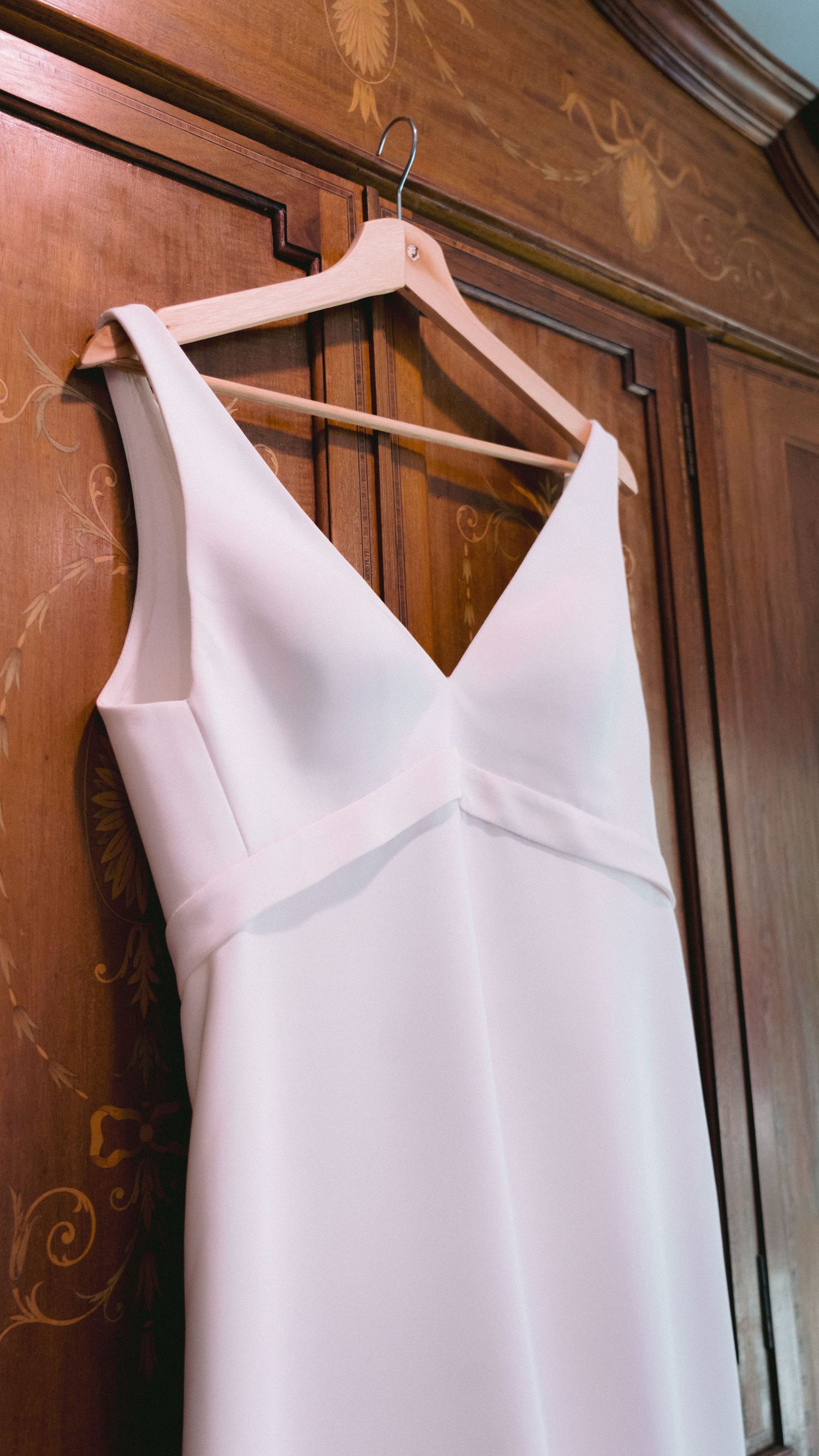 White wedding dresses hanging on wooden hangers against an ornate wooden wardrobe.