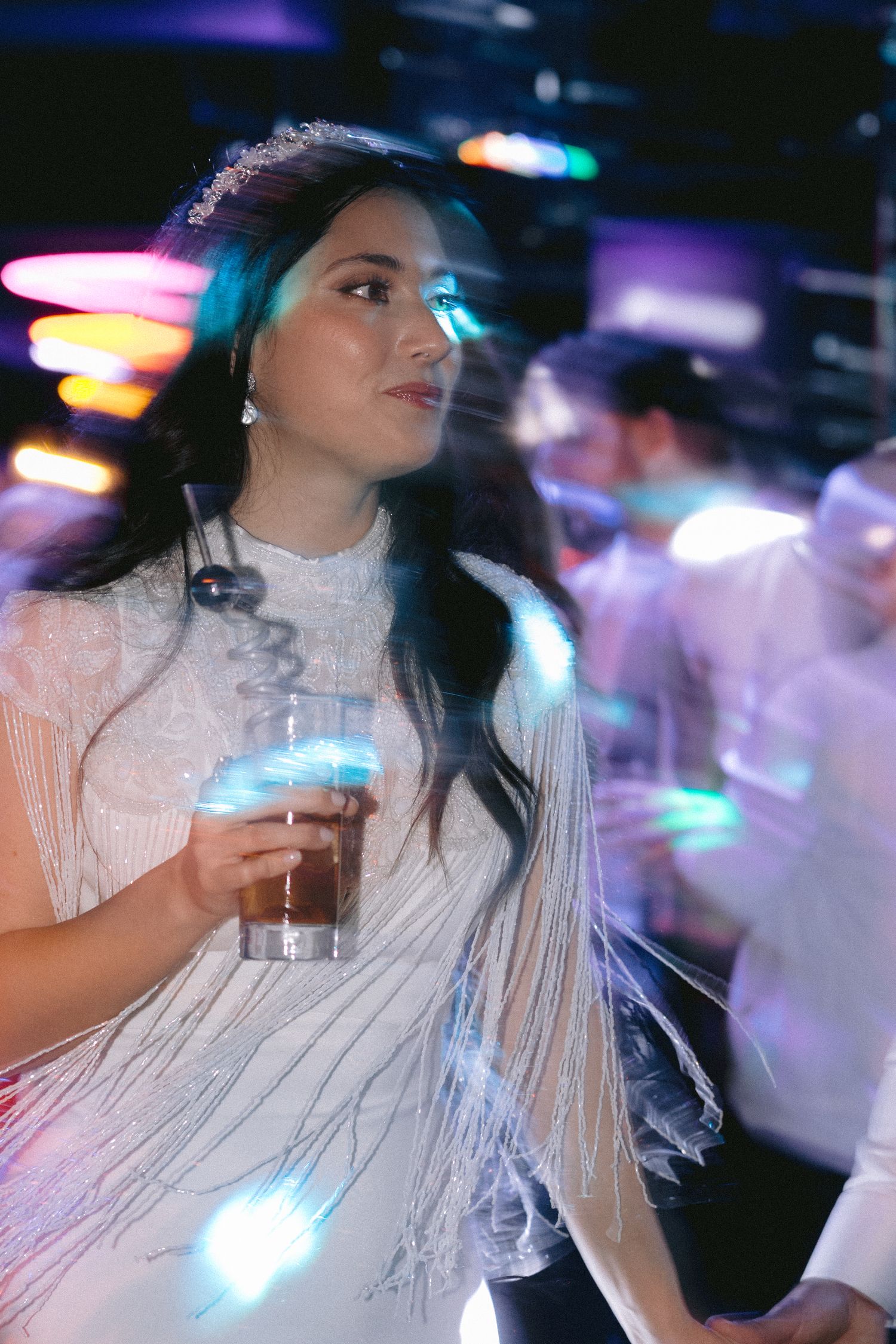 A guest in a fringed white dress enjoys drinks under vibrant nightclub lighting.