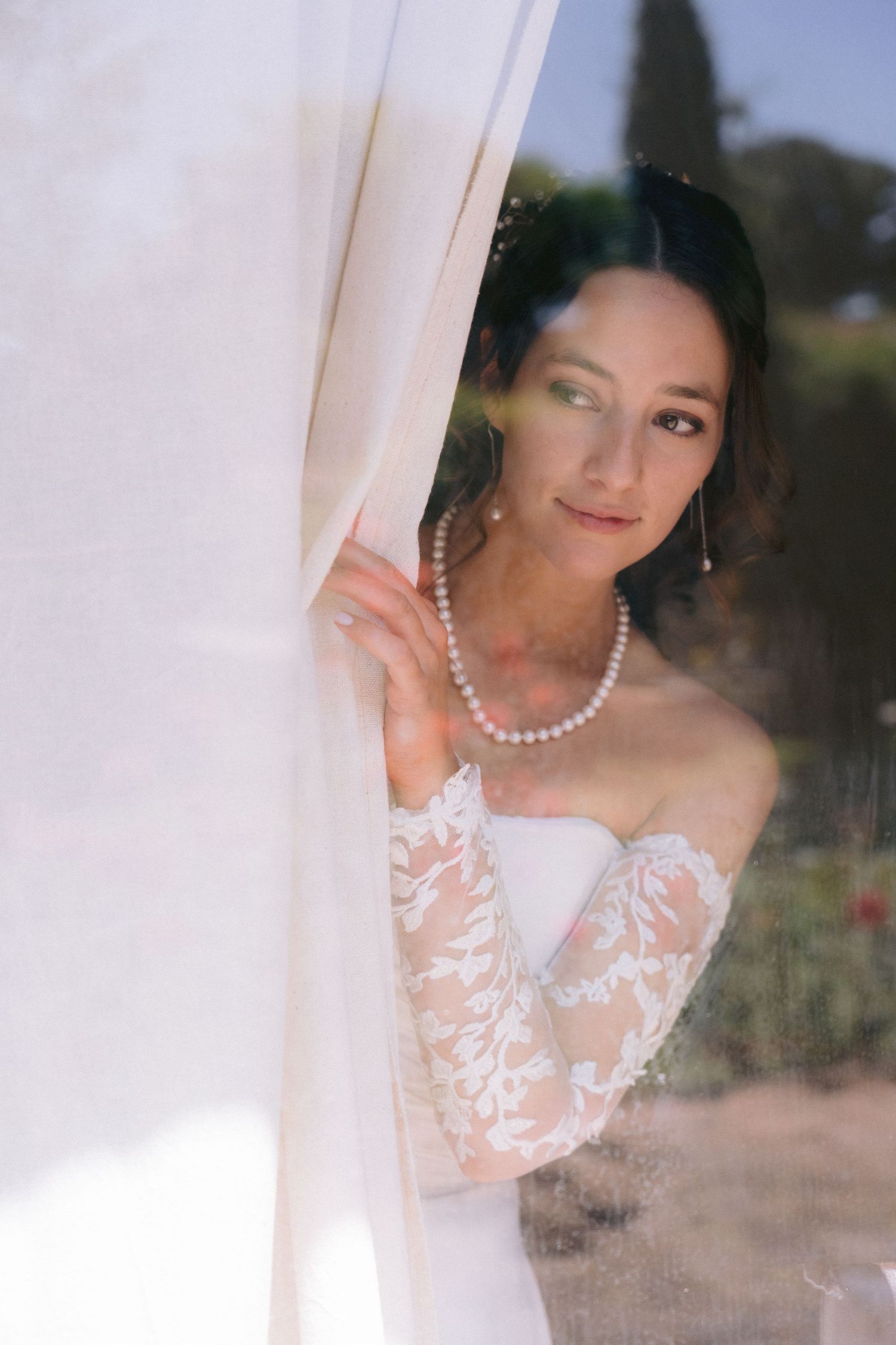 Elegant bride in white lace gown with pearl necklace poses behind sheer curtain in romantic outdoor setting.