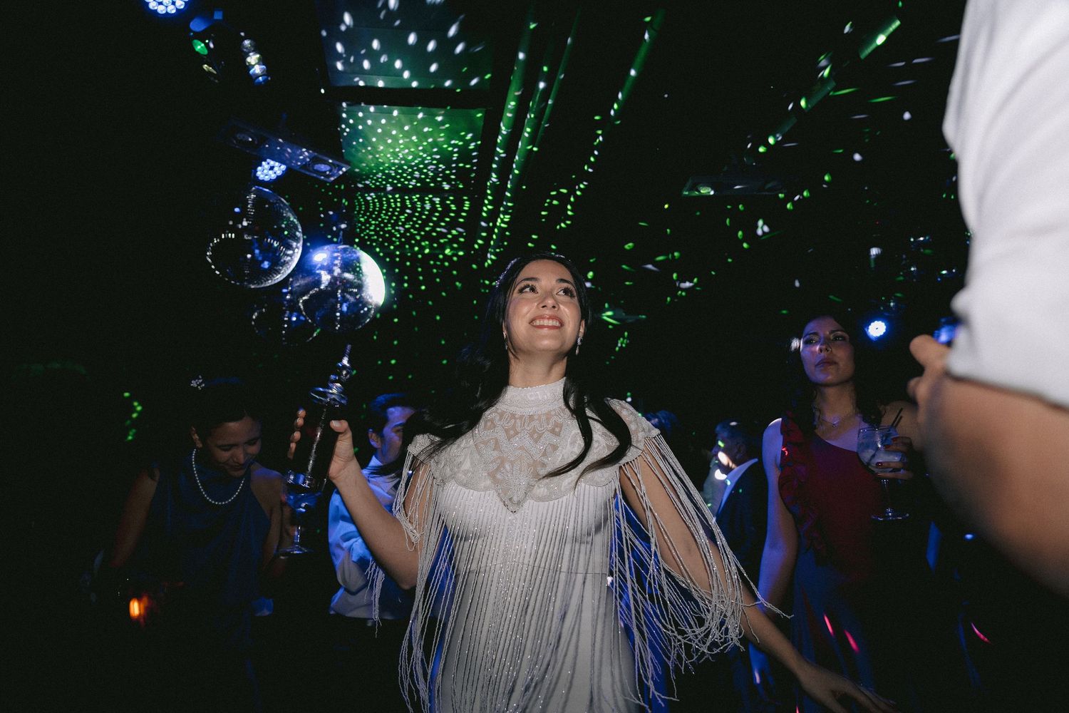 People dance and celebrate under blue and green disco lights at a nighttime party.