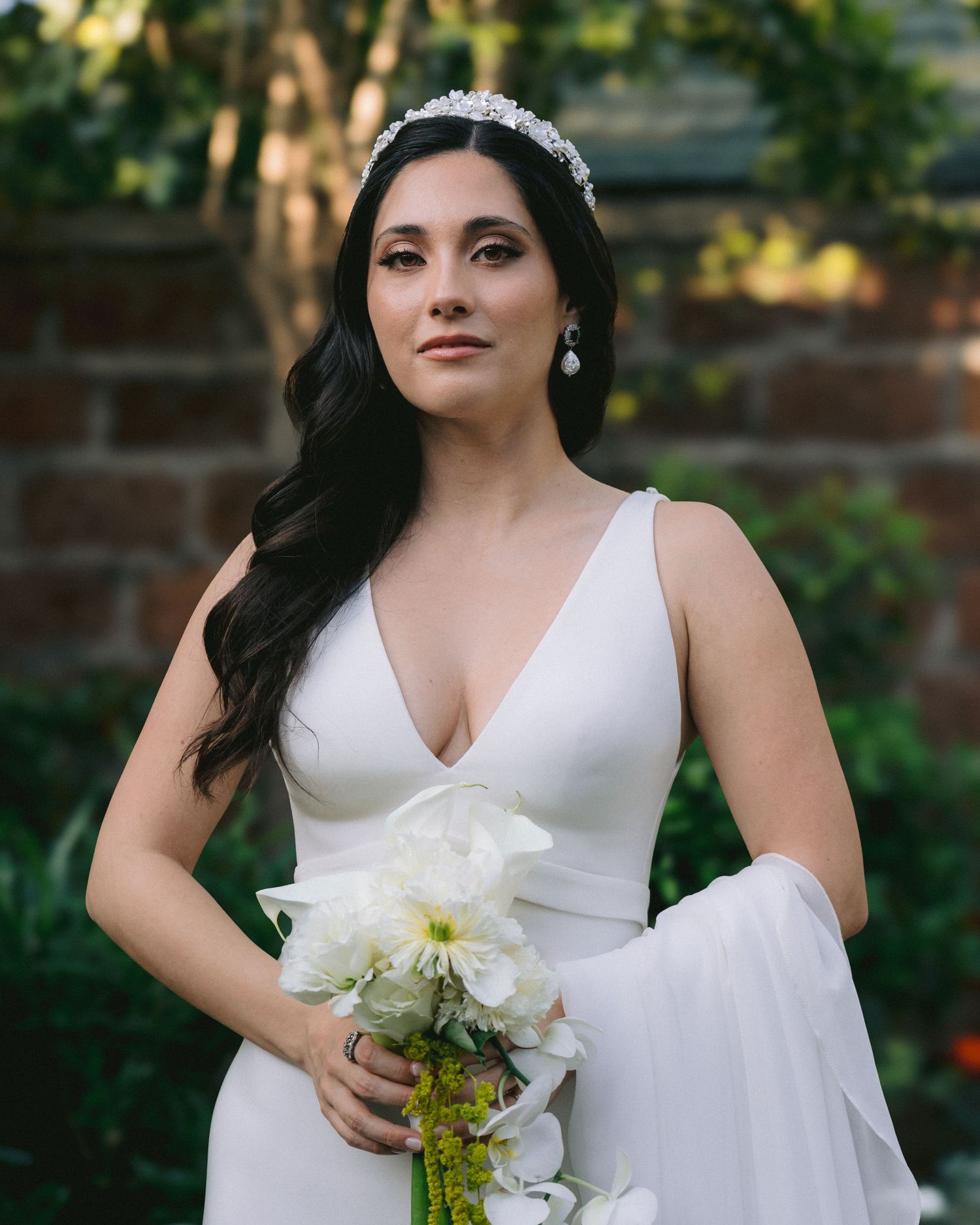 A bride wearing a sleek white gown and tiara stands in front of a brick wall in a garden setting.
