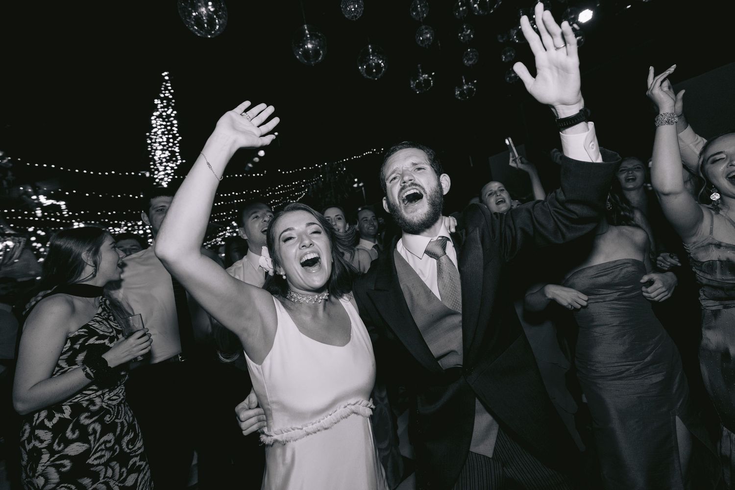 Energetic party crowd dancing with raised hands under dramatic dark lighting.