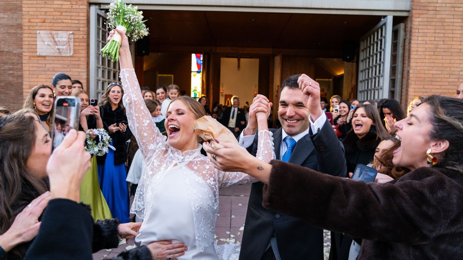 Newlyweds celebrate with joyful expressions while exiting a brick church surrounded by cheering guests.