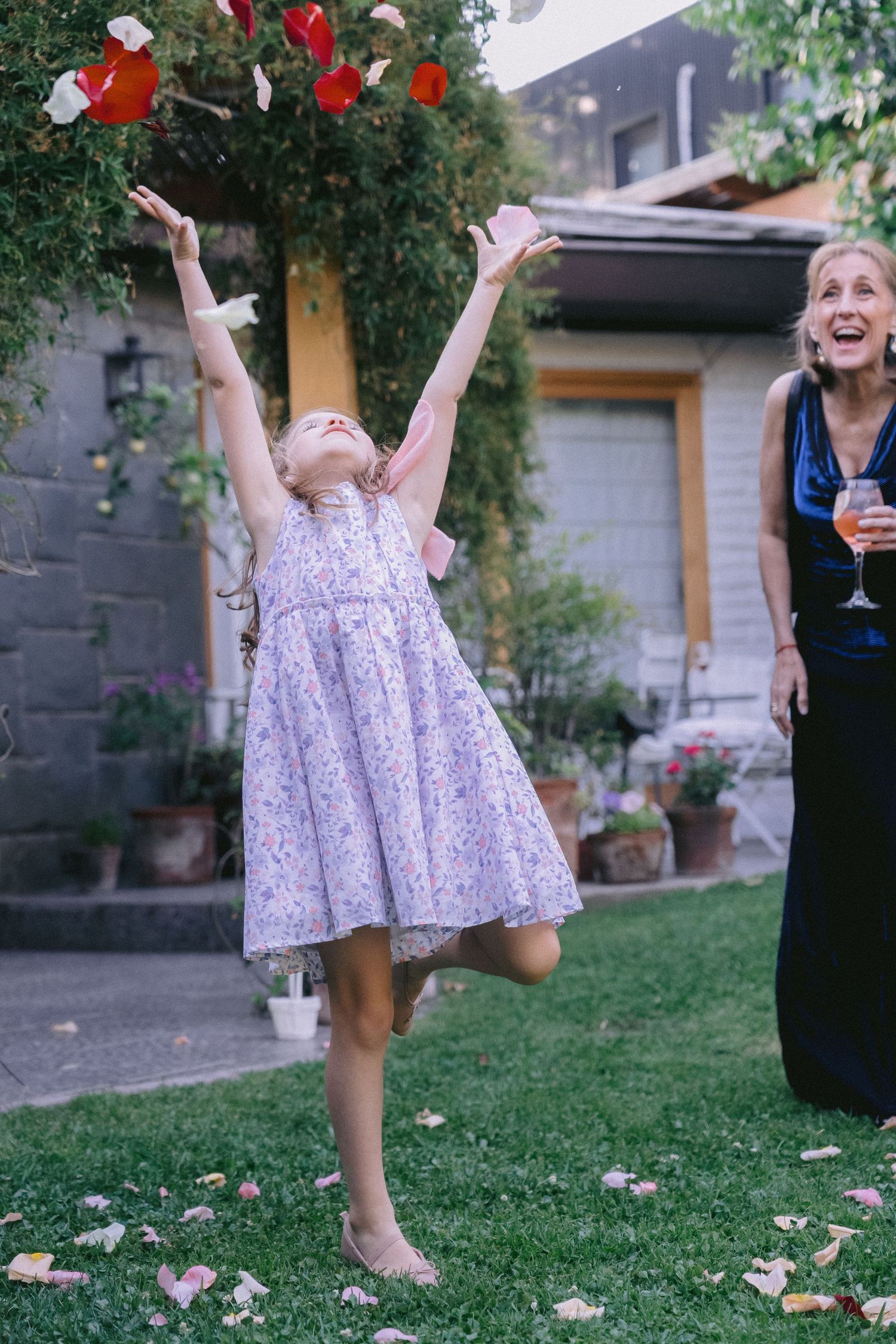 A child in a floral dress jumps joyfully on a lawn while playing with red balloons at an outdoor celebration.