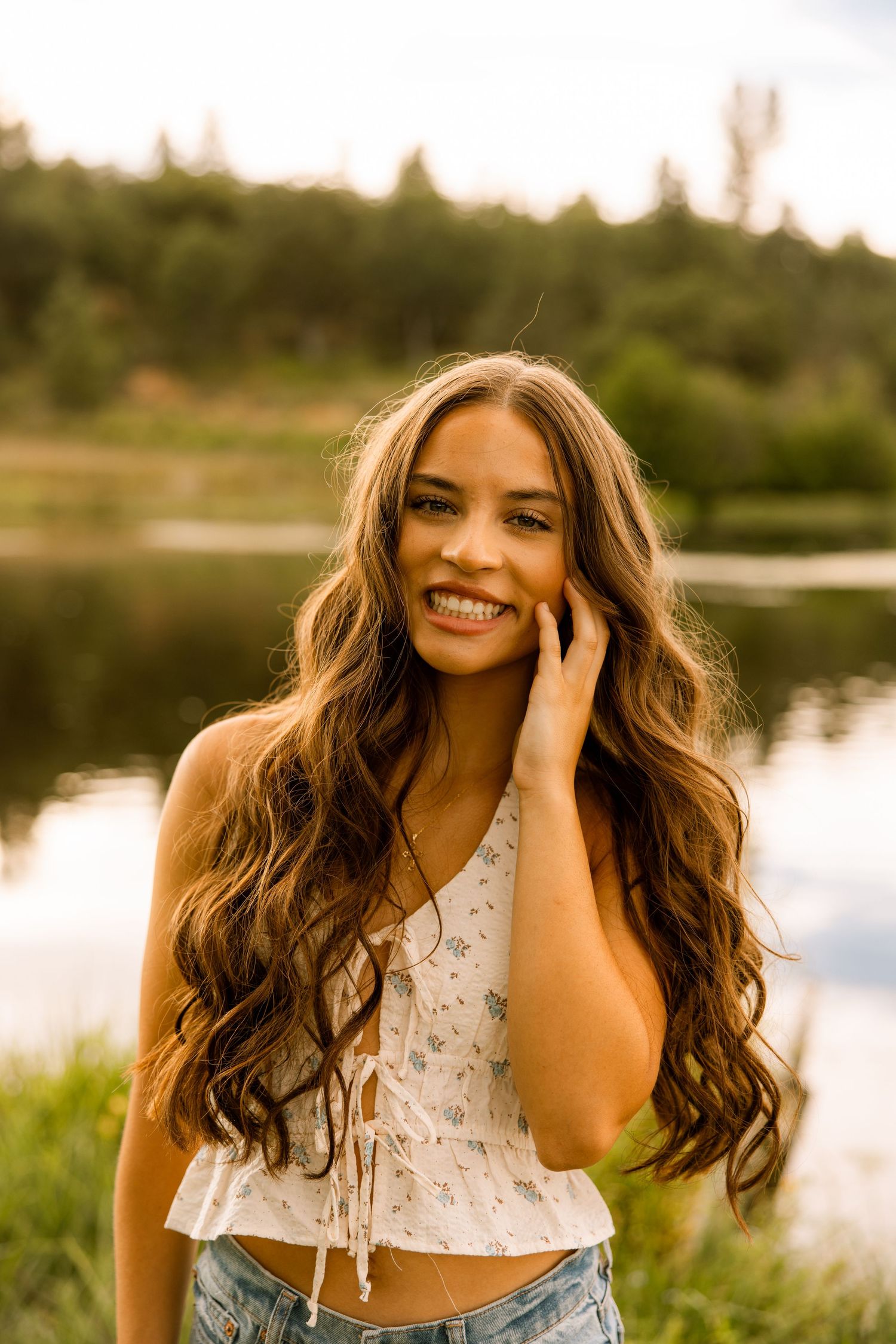 A person with wavy hair poses by a lake at sunset wearing a floral top.