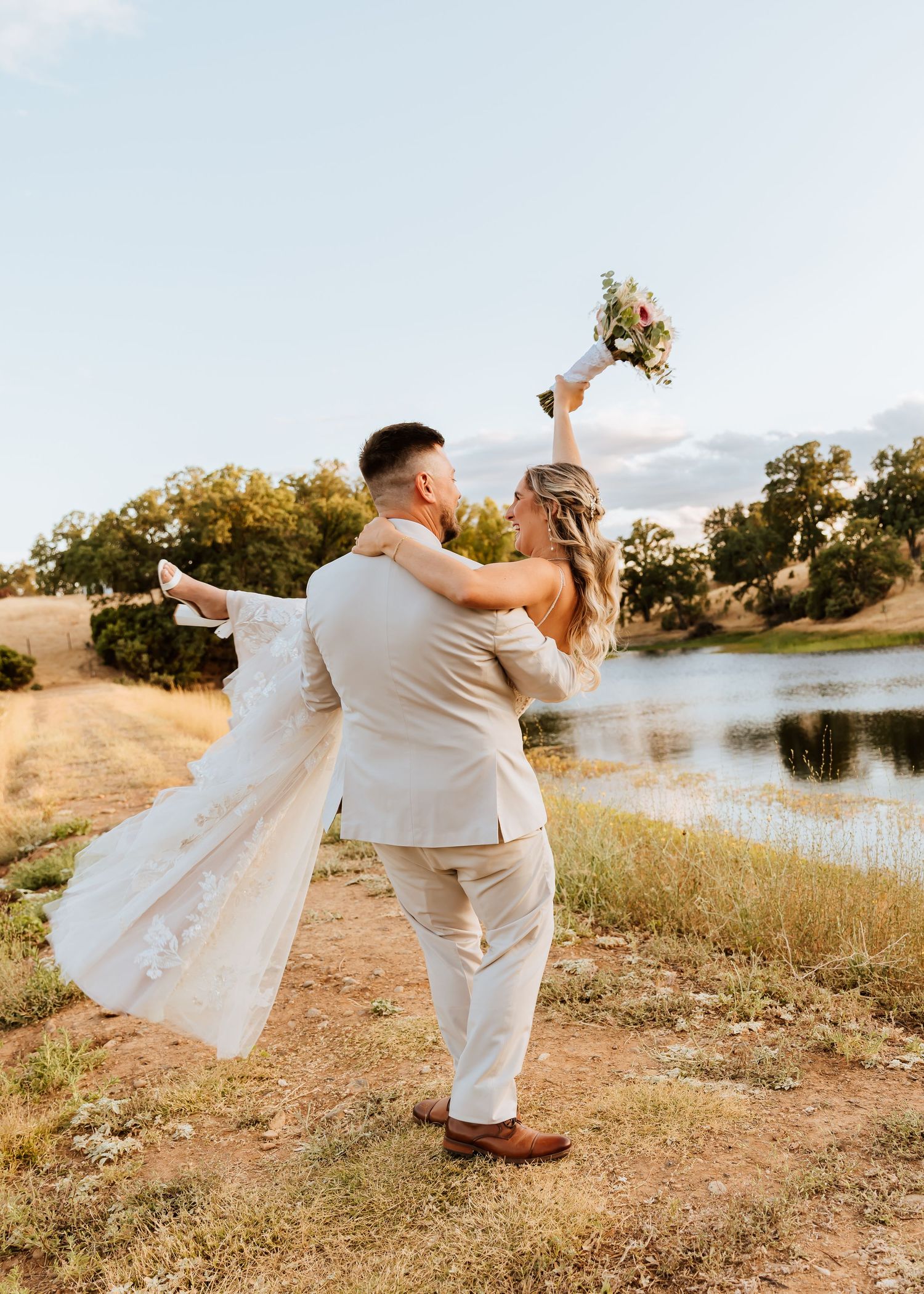 A playful wedding couple dancing and twirling outdoors near a serene lake at sunset.