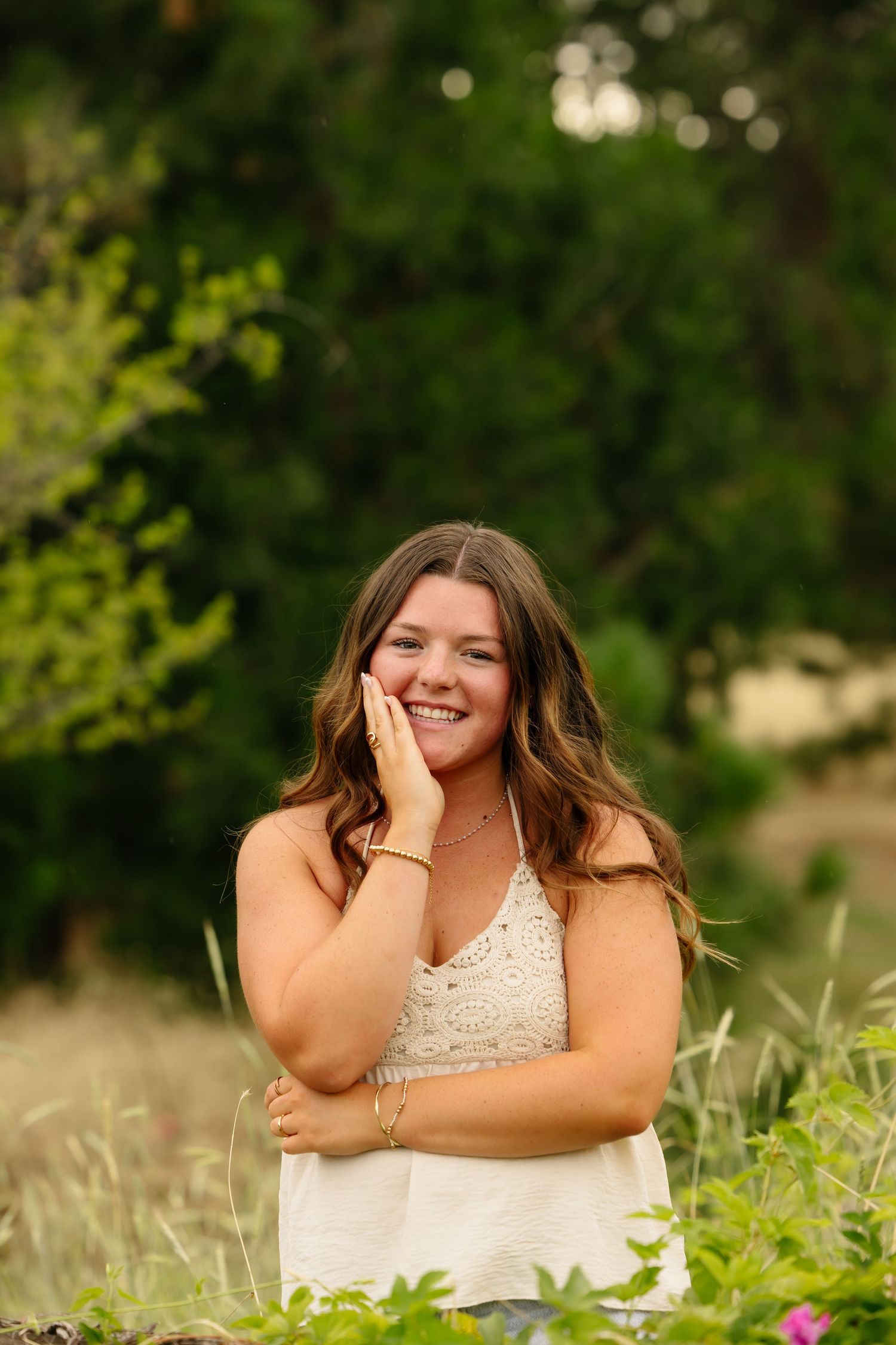 Smiling young woman with long brown hair in white lace tank top posing outdoors among green foliage.